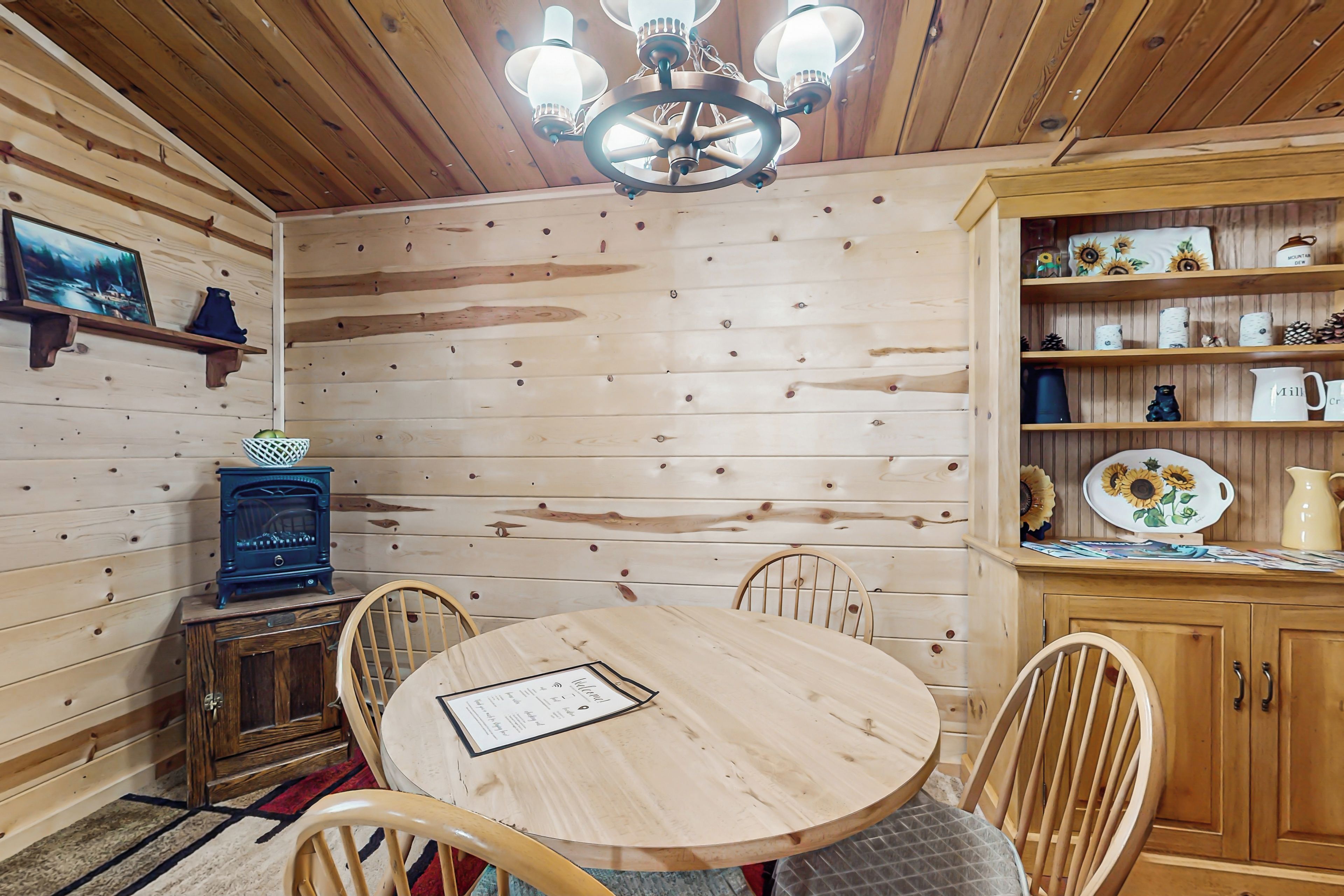 Interior de madera con una mesa redonda, sillas, una lámpara de araña y estantes con objetos decorativos.