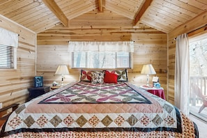 Un dormitorio rústico de cabaña de madera con una cama grande, dos lámparas de noche y un ventanal con persianas.
