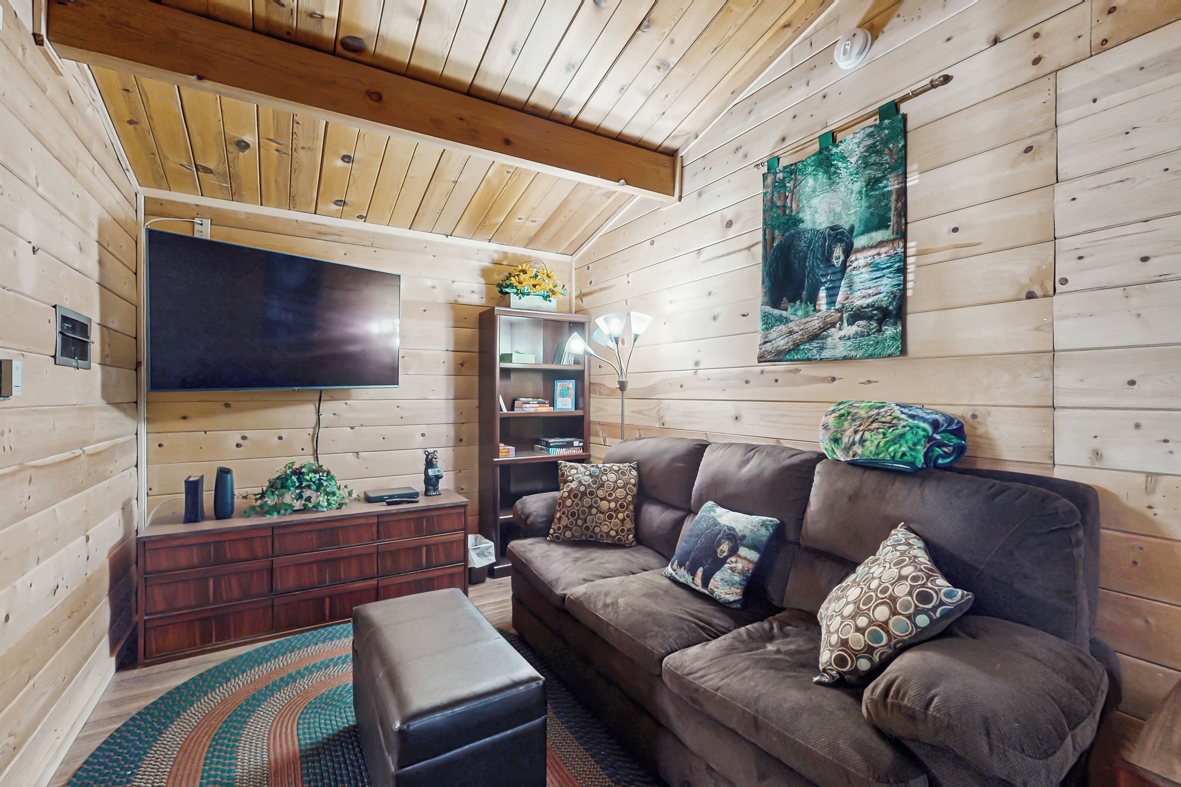 A cozy cabin interior with a wooden ceiling, a large flat-screen TV, a brown leather sofa with patterned cushions, a wooden cabinet, and a painting of a bear.