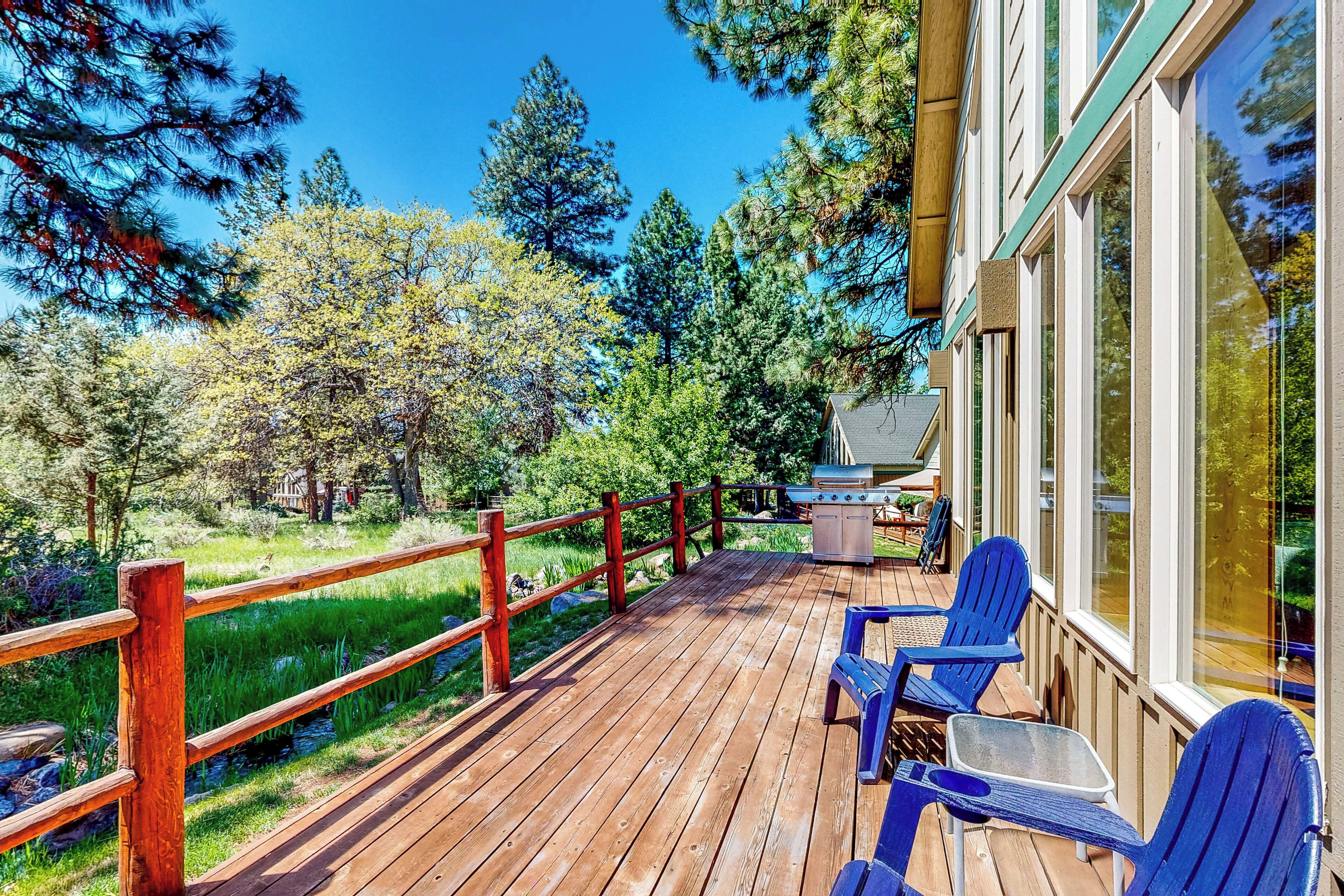 A wooden deck with a grill and blue chairs, surrounded by trees and a wooden fence.