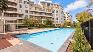 Una piscina con borde de baldosas, rodeada por un edificio residencial de varios pisos con balcones.
