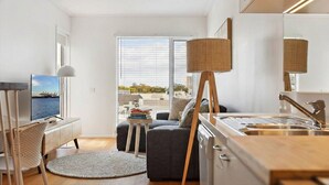 Una habitación moderna y luminosa con un ventanal grande, comedor, cocina con lavamanos y una pequeña mesita con libros.