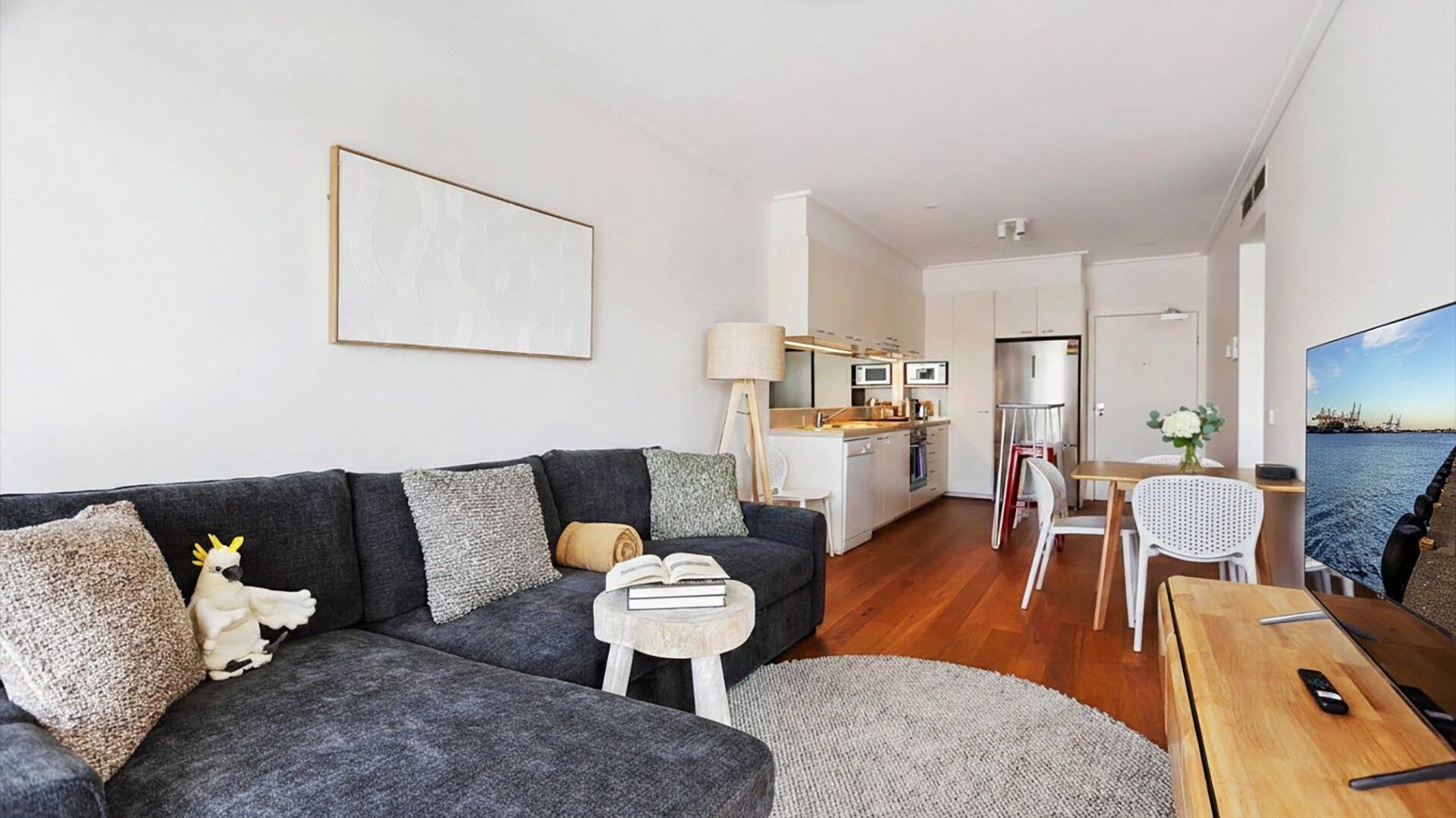 Un salon moderne avec un canapé gris, une petite table d’appoint et une table à manger en bois. Une cuisine avec des meubles blancs et une vue sur la mer est visible à travers une porte ouverte.