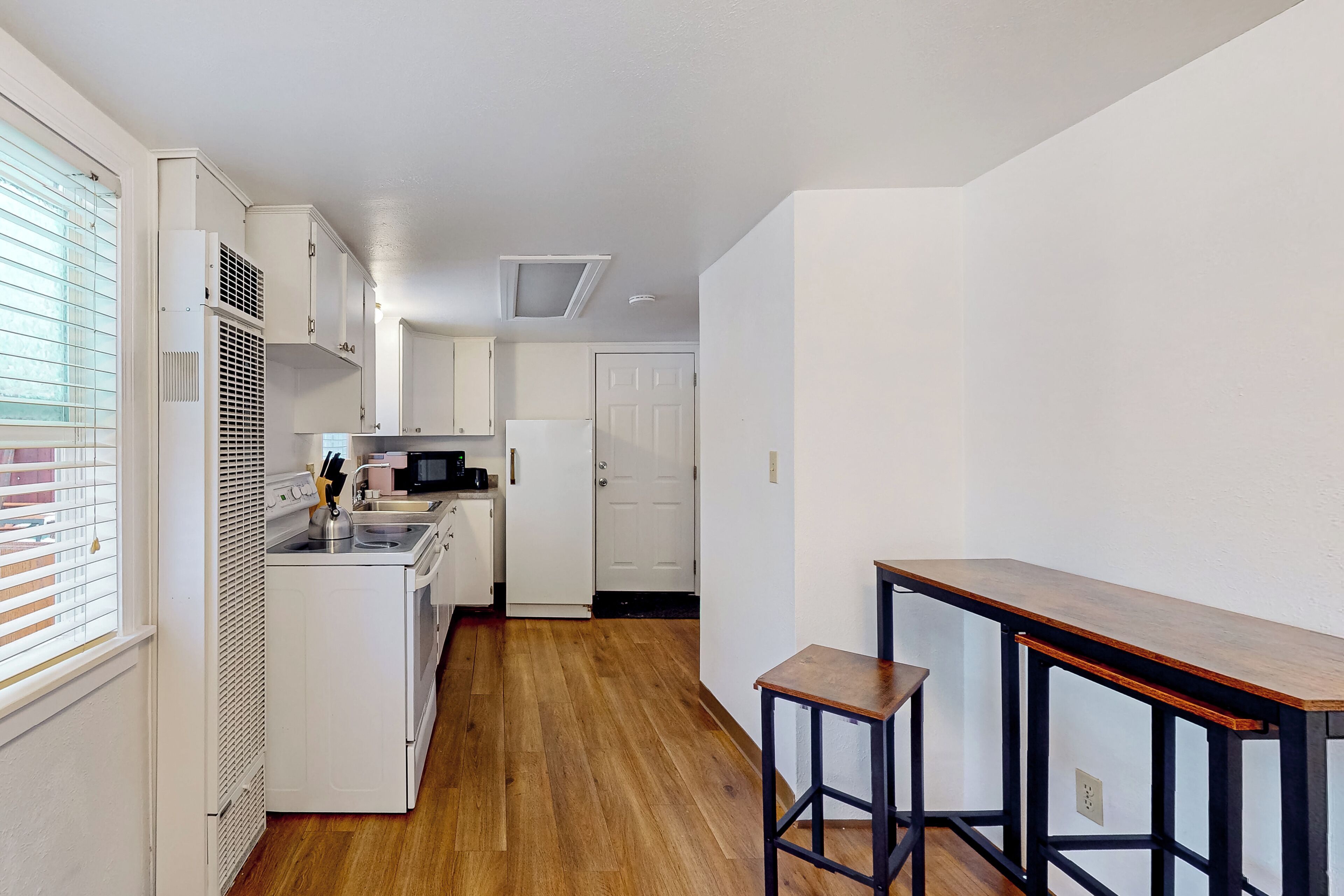 A compact kitchen with white cabinets, a microwave, and a stove. There is a wooden floor and a small wooden table with two stools.