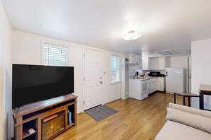 A modern living room with a flat-screen TV, wooden entertainment unit, and a beige sofa. A kitchen with white appliances and a small dining area is visible in the background. - 1BR Sunlit Retreat w/ Patio - Near Golf & Parks (Klamath Falls)