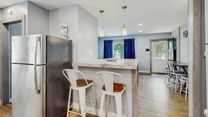 Une cuisine moderne équipée d’un réfrigérateur, de deux tabourets de bar, d’un plan de travail en marbre et de luminaires suspendus.