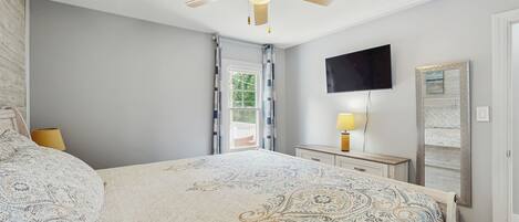 A bedroom with a large bed, a ceiling fan, a TV, a mirror, and a window with curtains.