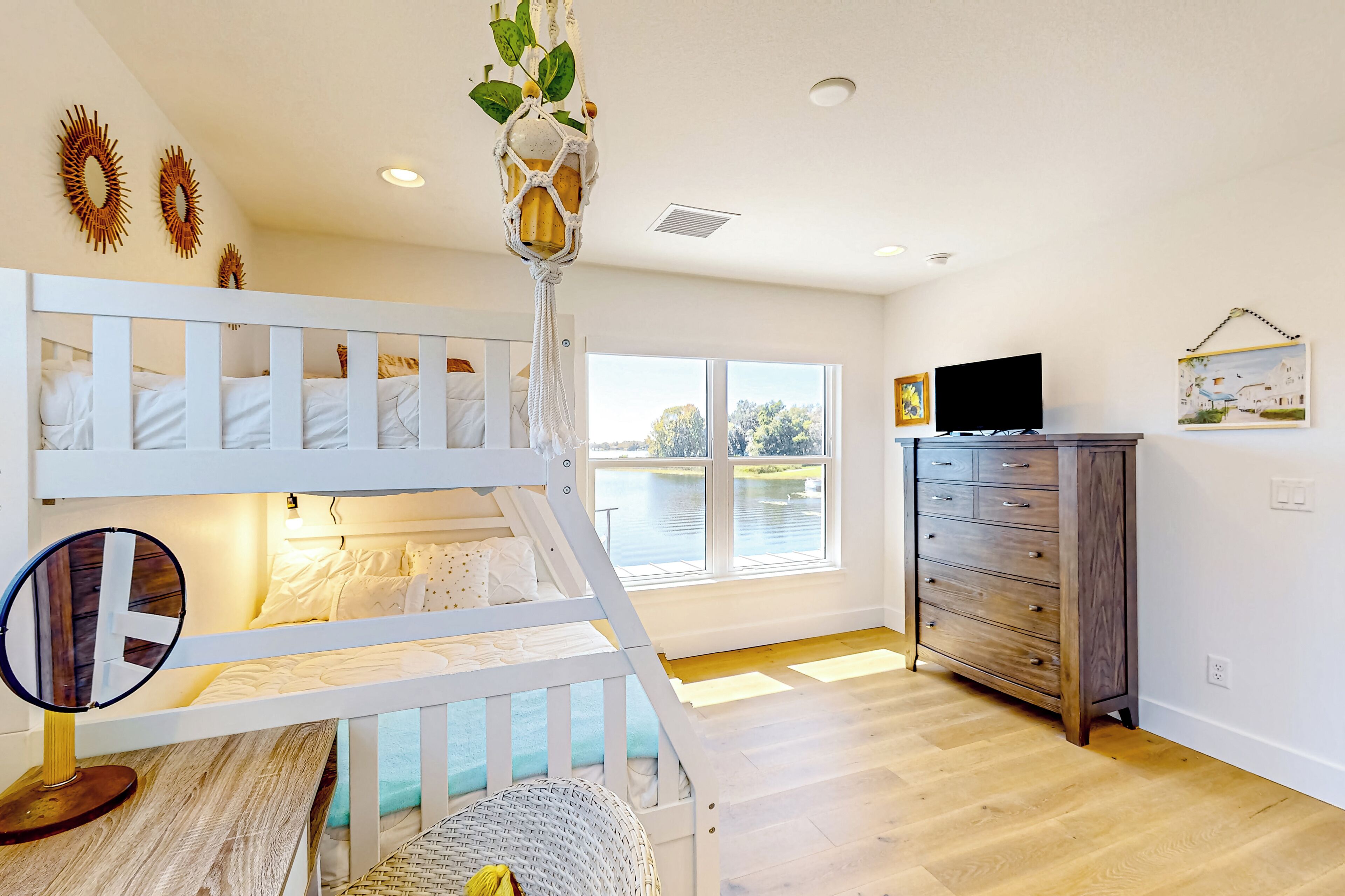 A room with a bunk bed, a vanity with a mirror, a dresser, and a large window with a view of a body of water.