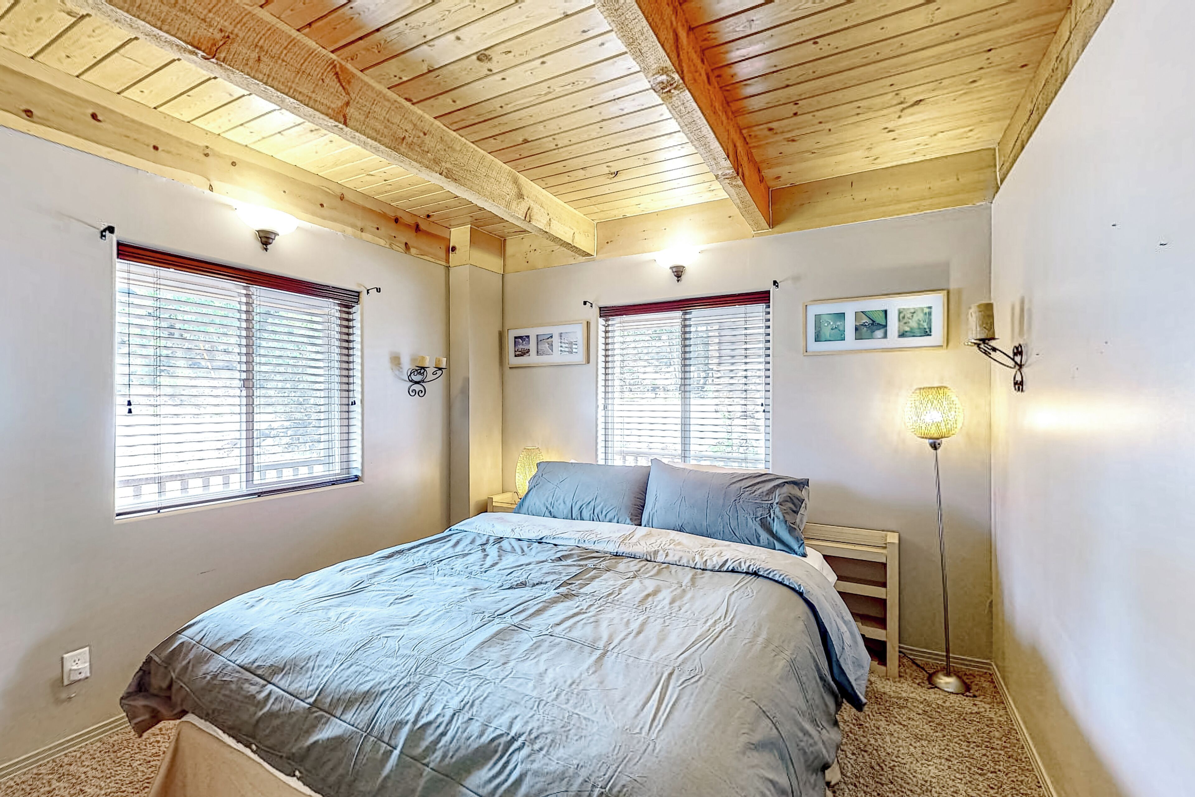 A bedroom with a wooden ceiling, two windows with blinds, a bed with a grey bedspread, a bedside lamp, and a framed picture on the wall.