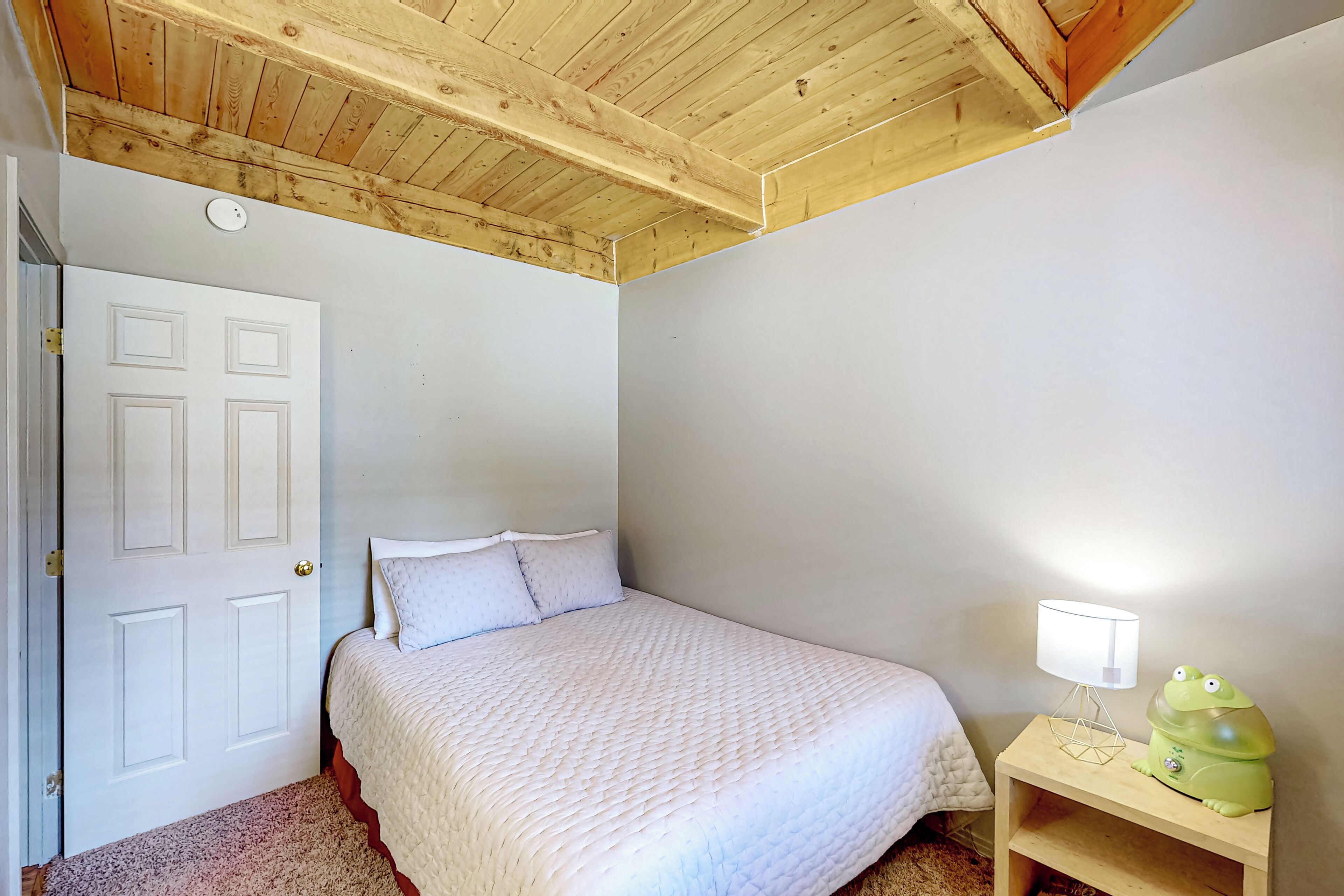 Une chambre avec un plafond en bois, un lit recouvert d’une couette blanche, une table de chevet avec une lampe en forme de grenouille verte et une porte blanche.