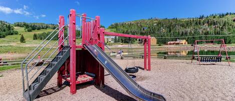 Un parque infantil con un tobogán y una estructura de escalada, rodeado de grava y una vista panorámica de colinas y árboles.