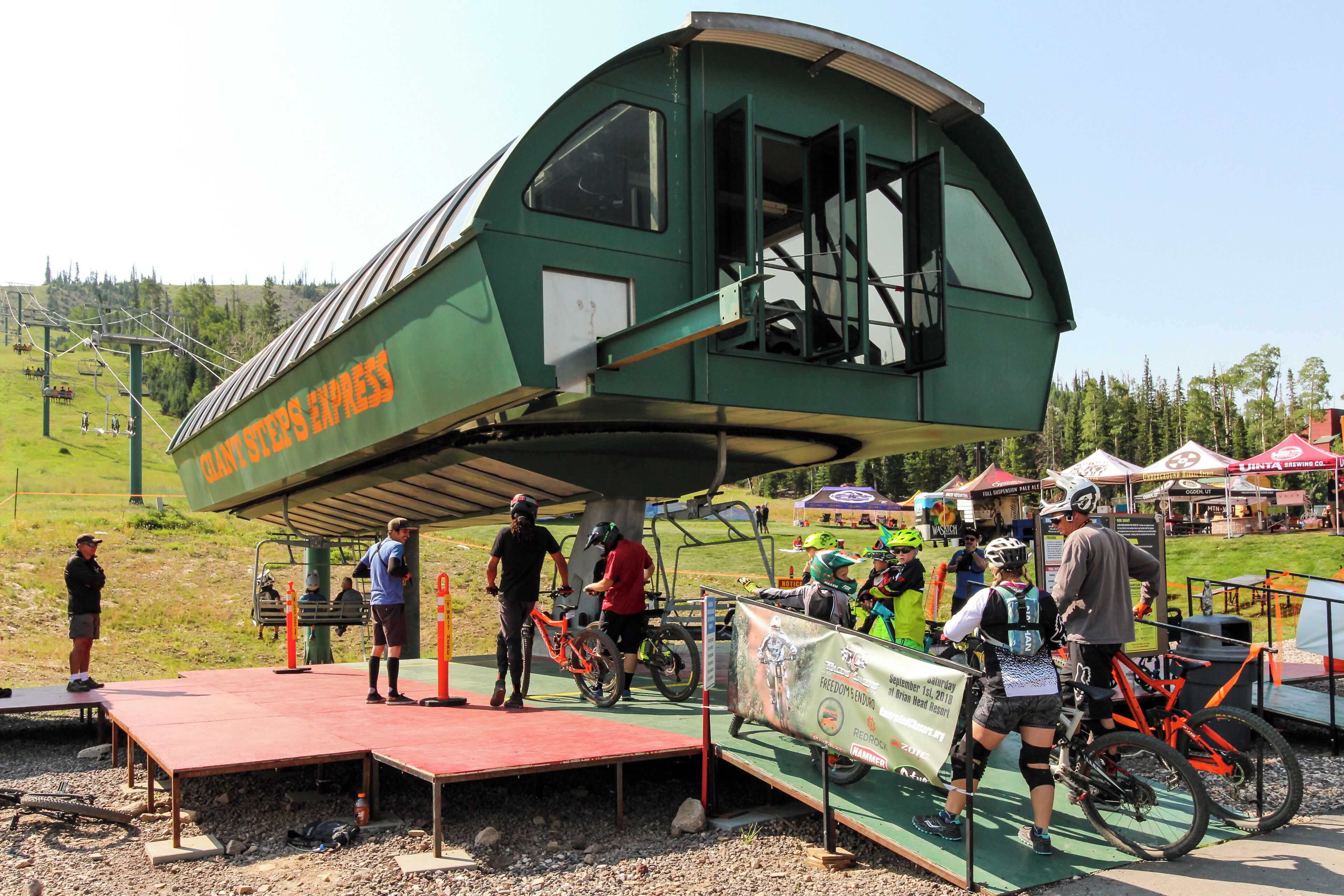 A green, elevated viewing platform with a curved roof, surrounded by a green area with trees and a ski lift in the background.