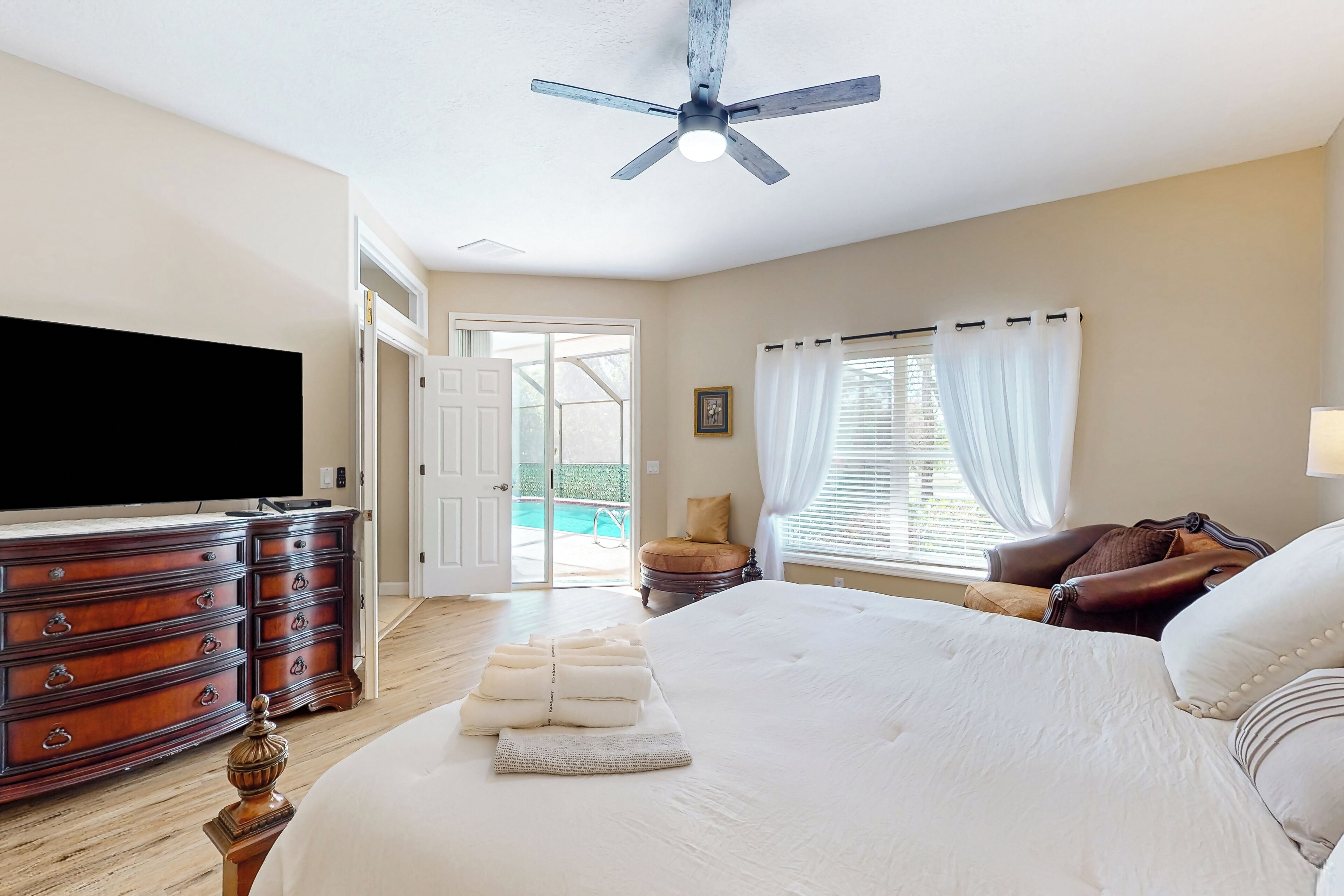A bedroom with a large bed, a wooden dresser, a ceiling fan, and a view of the outdoors.