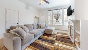 A modern living room with a sectional sofa, a patterned rug, a ceiling fan, and a fireplace. - 3BR Mountain View Home w/ Patio & Streaming (Pocatello)