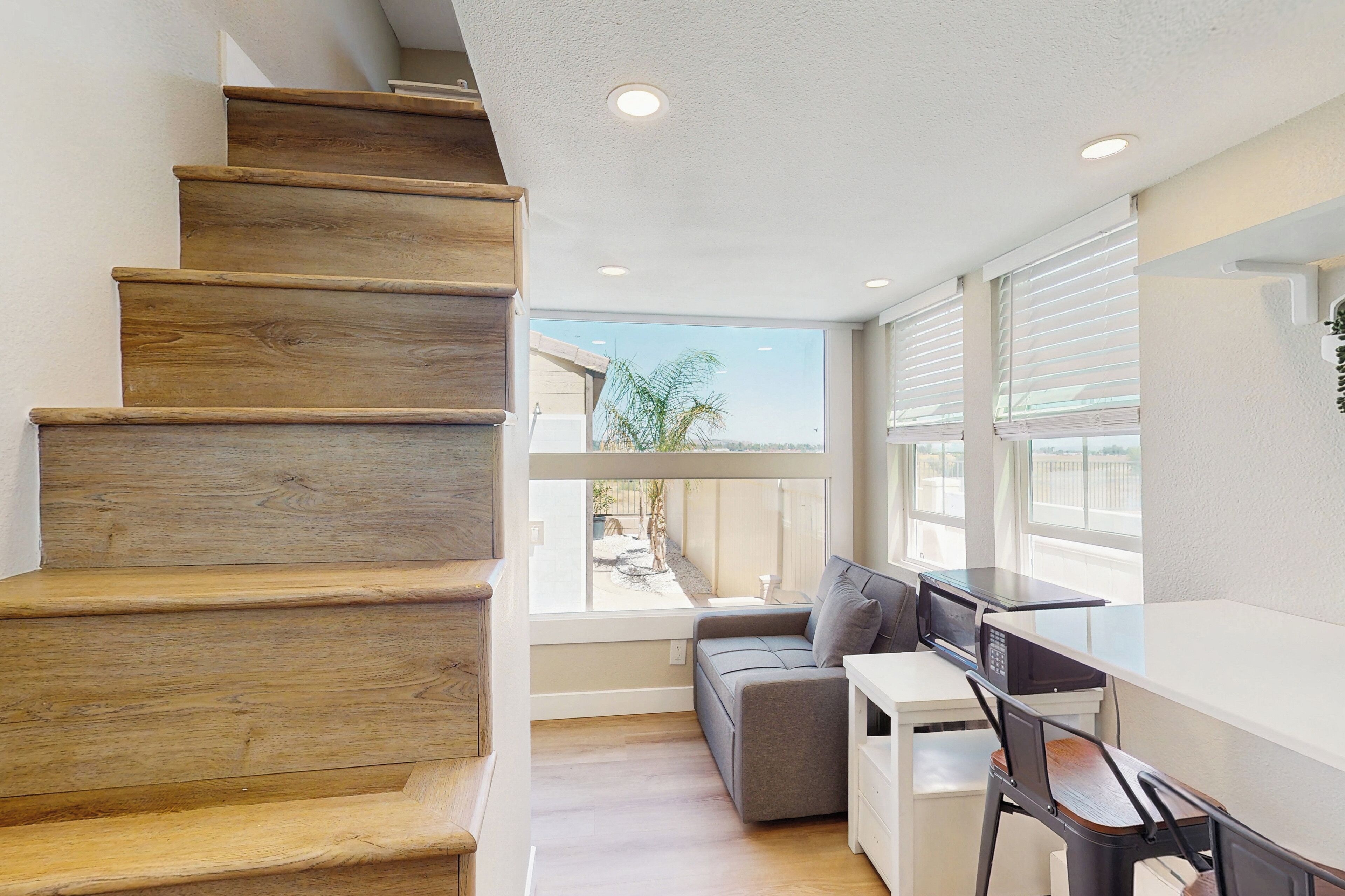 A modern interior with a wooden staircase, a gray sofa, a white desk, and large windows offering a view of the outdoors.