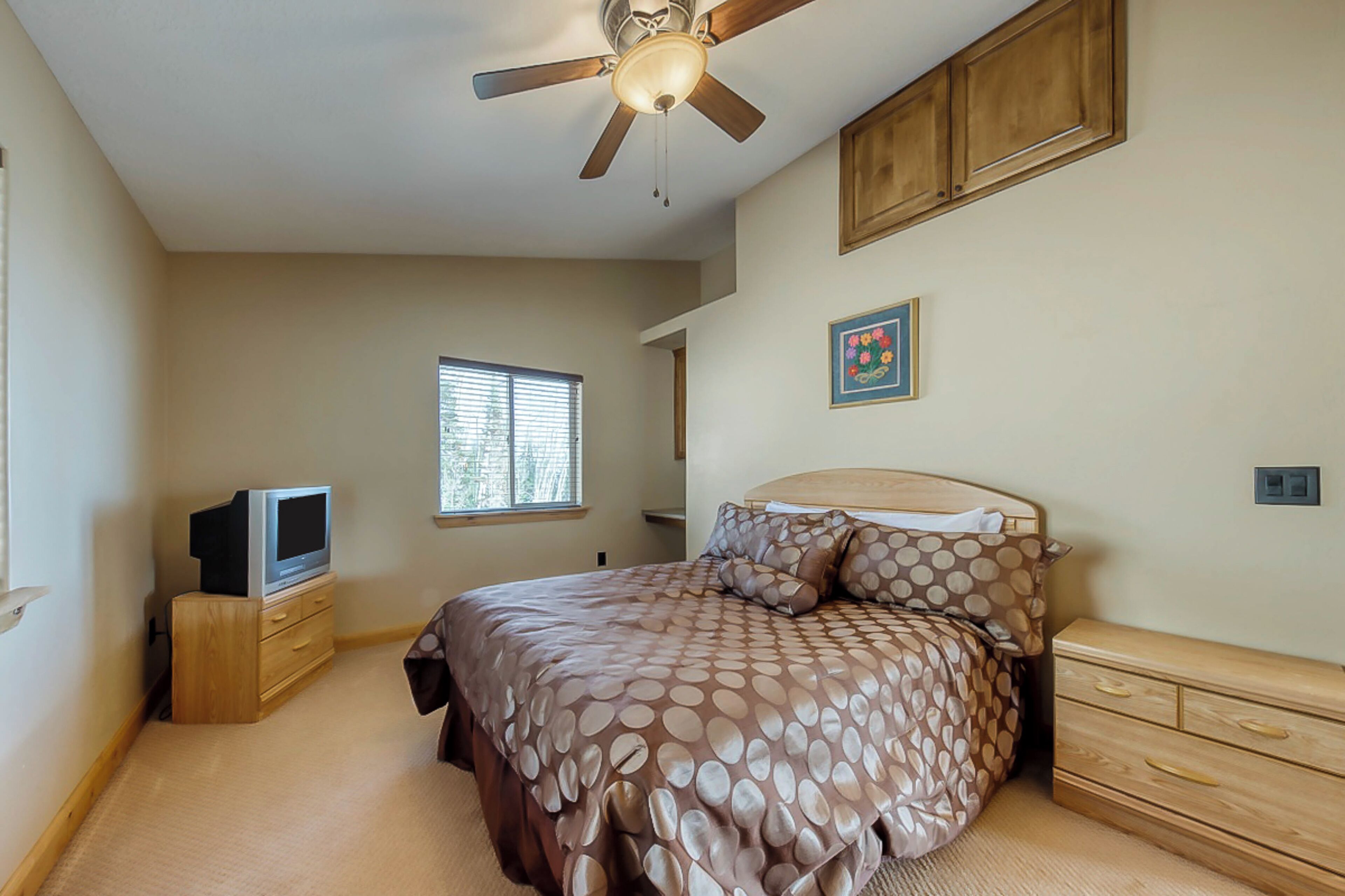A bedroom with a bed, a ceiling fan, a television on a wooden stand, a window, and a painting on the wall.