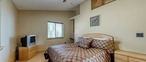 A bedroom with a bed, a ceiling fan, a television on a wooden stand, a window, and a painting on the wall.
