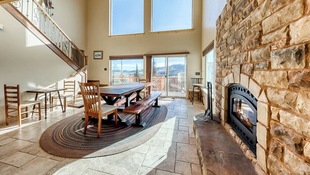 A spacious living area with a stone fireplace, a dining table with chairs, a staircase, and large windows offering a view of the outdoors. - Brian Head 'view-topia' w/ Mtn Views & King Suite (Brian Head)
