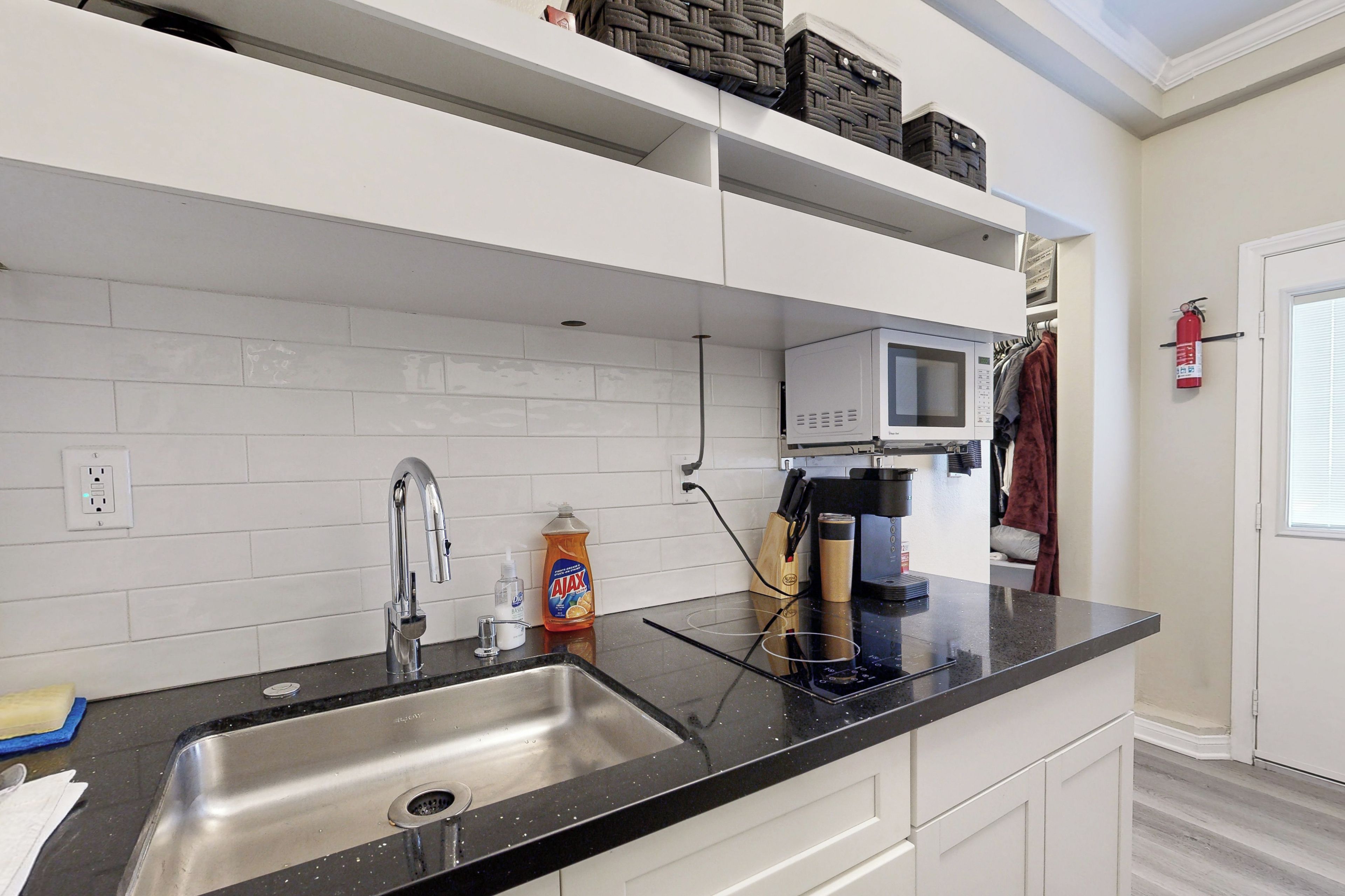 Una cocina moderna con encimera negra, pileta de acero inoxidable, muebles blancos, microondas y extintor.