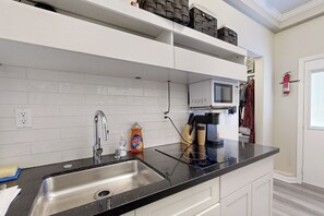 Una cocina moderna con encimera negra, pileta de acero inoxidable, muebles blancos, microondas y extintor.