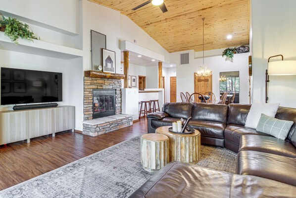 A modern living room with a leather sofa, a fireplace, a television, and a dining area in the background. - 3BR Home w/ Hot Tub, Grill & Wraparound Deck (Show Low)