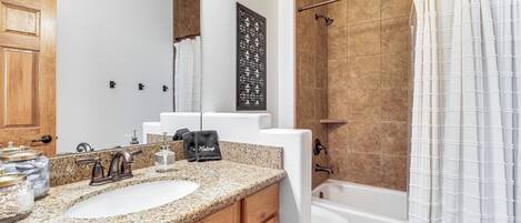 Un baño con encimera de granito, tocador de madera, lavabo blanco, espejo, cortina de ducha y bañera.