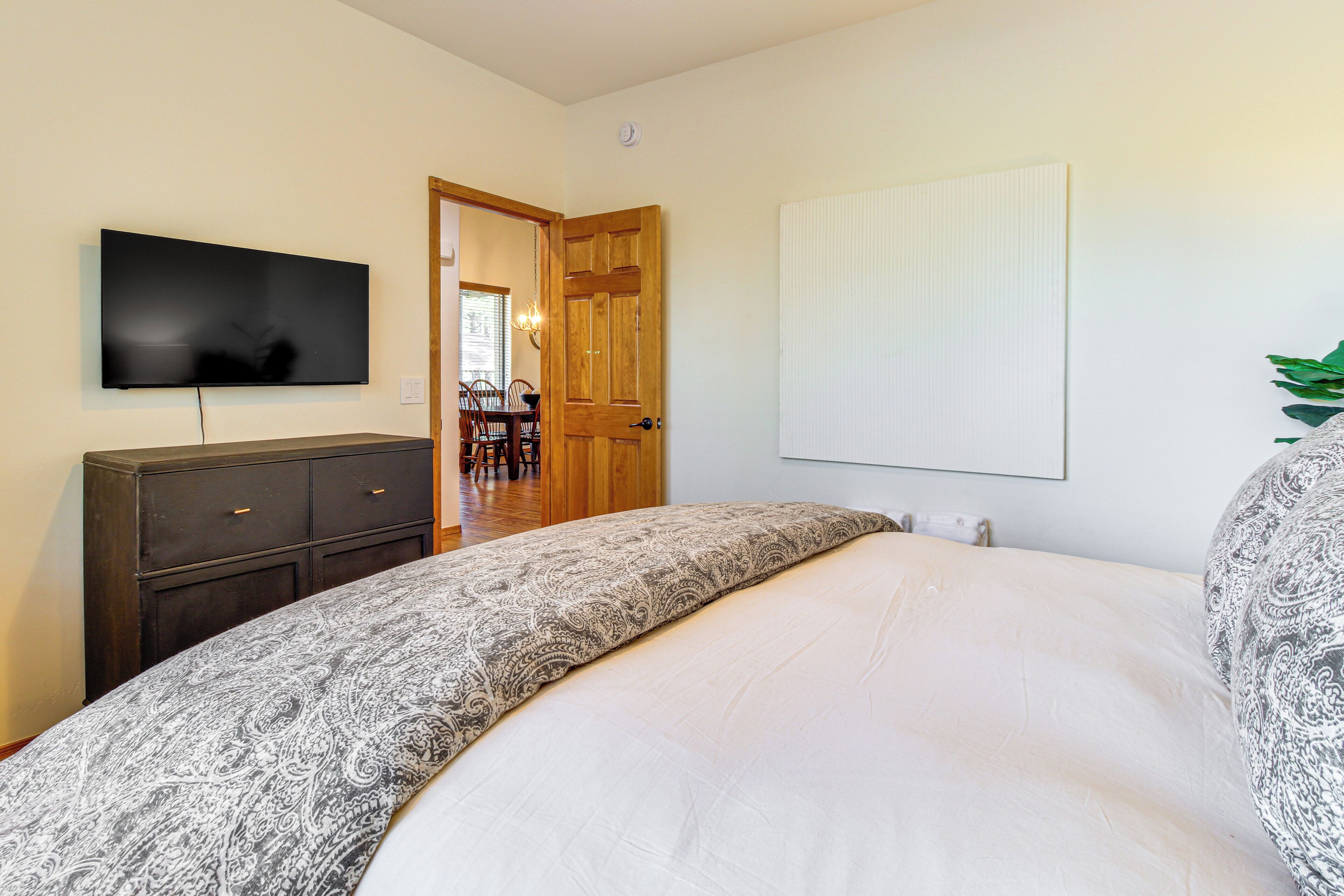 A bedroom with a bed, a TV mounted on the wall, a wooden door, and a dining area visible through the door.