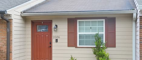 A single-story house with a brown front door, a window with shutters, and a small tree in the front yard.