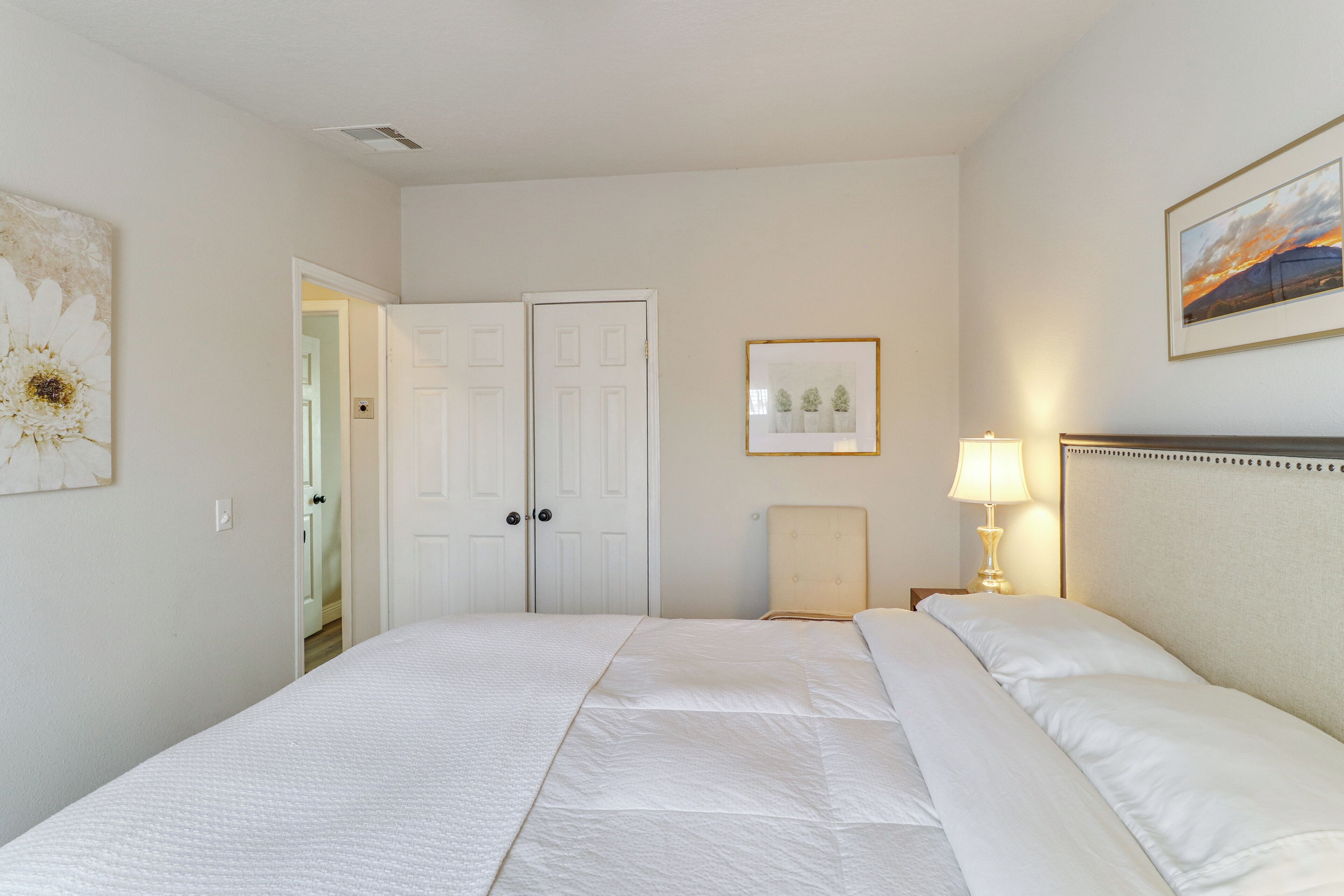A hotel room with a large bed, a nightstand with a lamp, a framed picture on the wall, and a door leading to another room.