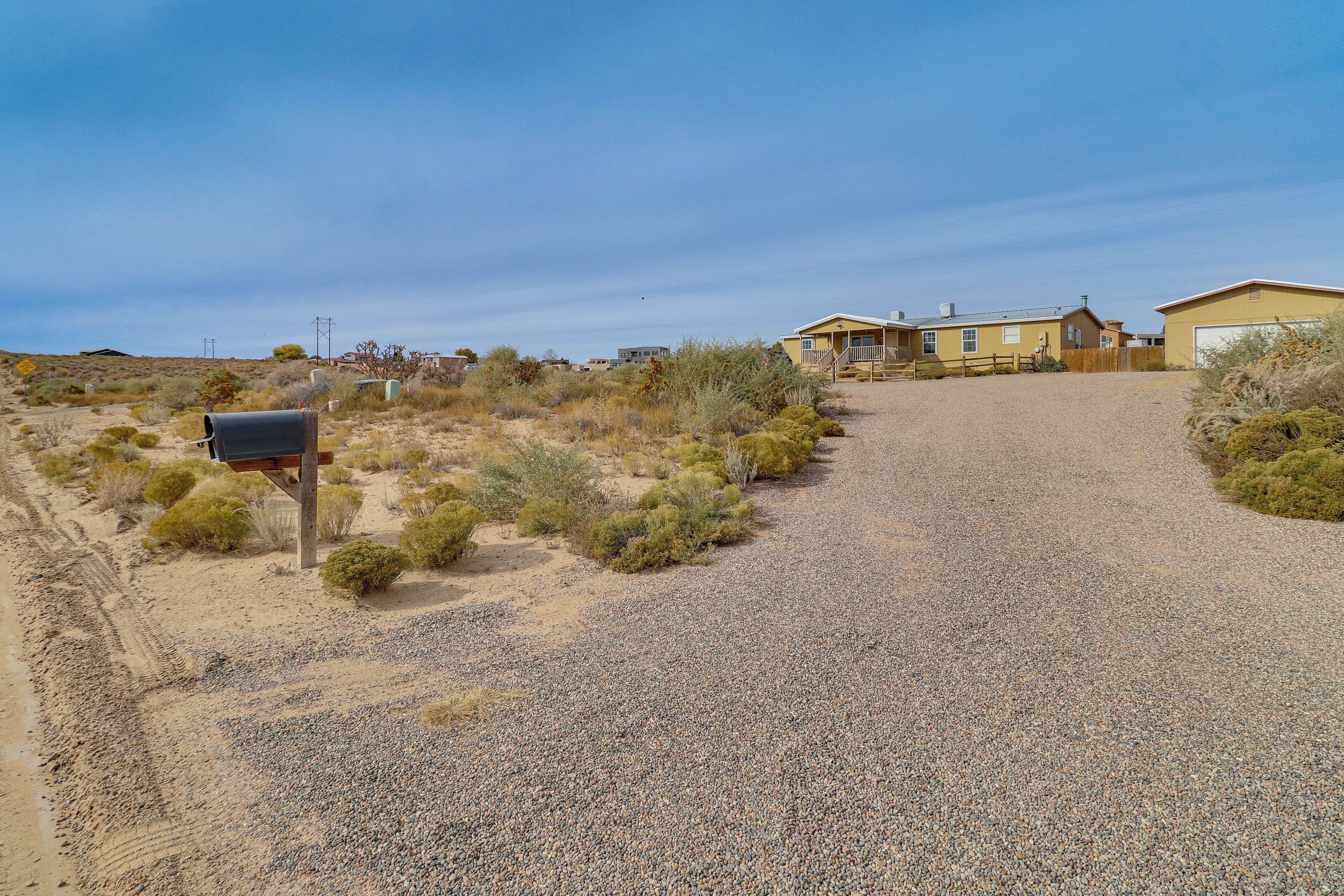 A gravel driveway leading to a yellow house with a garage, surrounded by sparse vegetation and a clear sky.