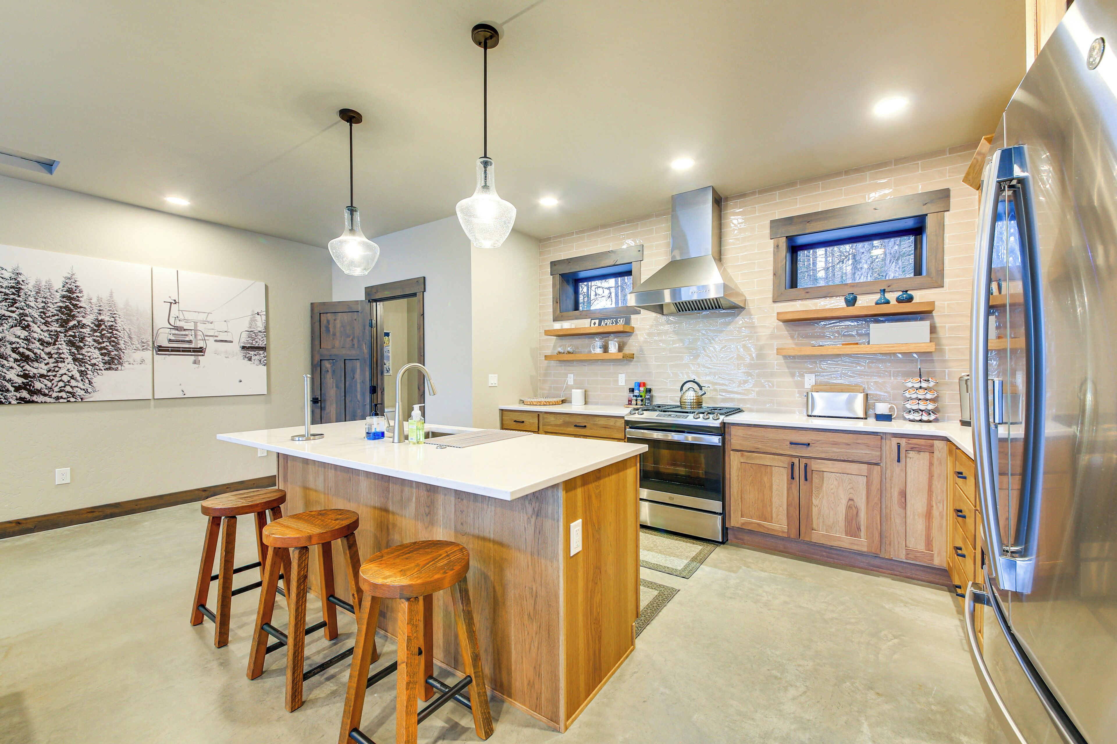 A modern kitchen with a central island, wooden stools, stainless steel appliances, and a window with a view of trees.