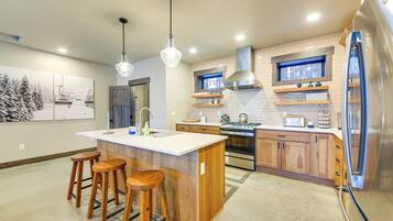 A modern kitchen with a central island, wooden stools, stainless steel appliances, and a window with a view of trees.