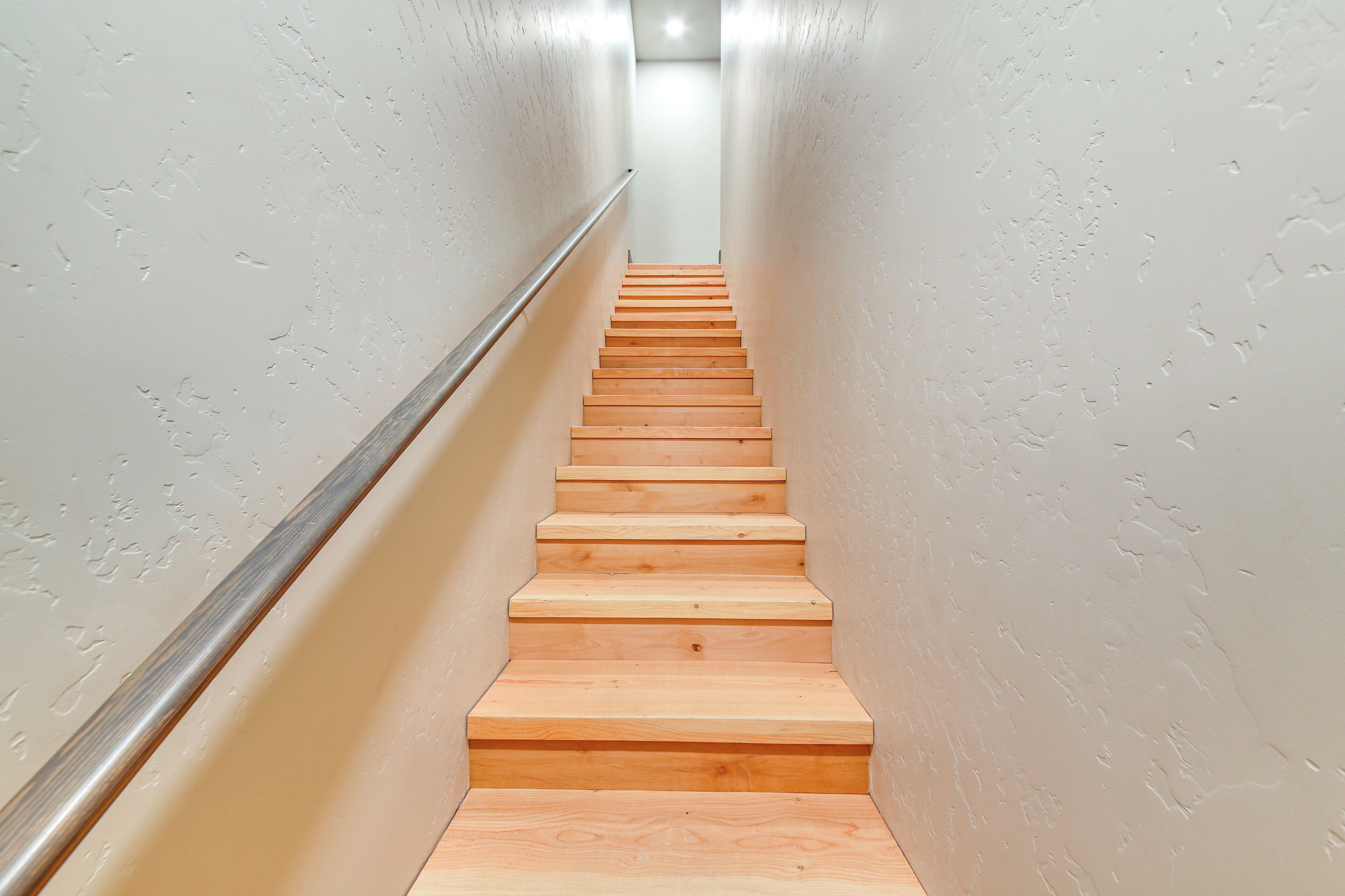 A wooden staircase with a metal handrail, leading upwards in a narrow hallway with plain walls.