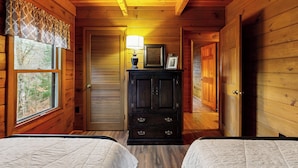 Una habitación de cabaña de madera con dos camas, un aparador, una lámpara y una puerta que da a otra habitación.