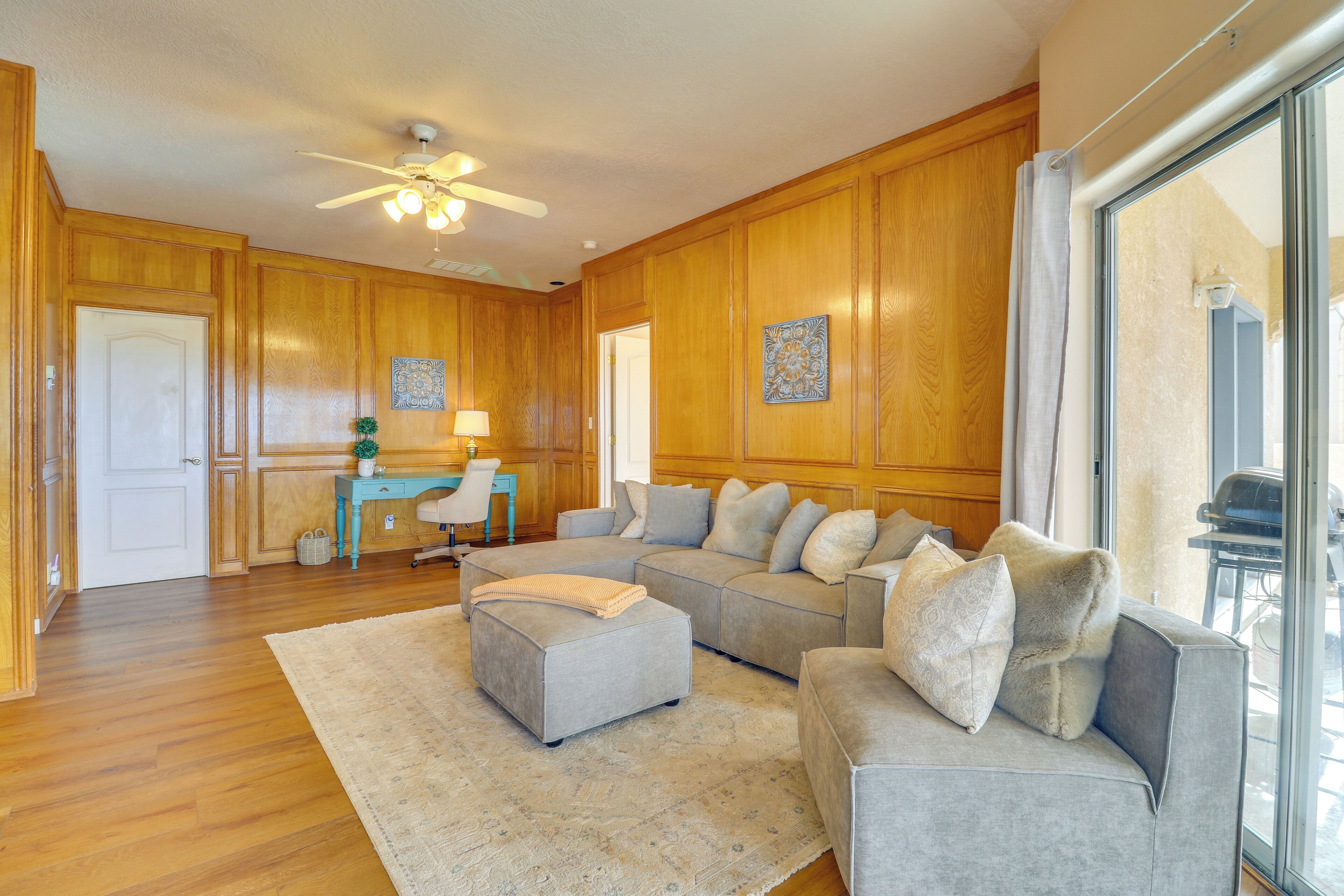 A spacious living room with a large gray sectional sofa, a matching ottoman, and a wooden floor. There is a ceiling fan, a window with curtains, and a small desk with a lamp.