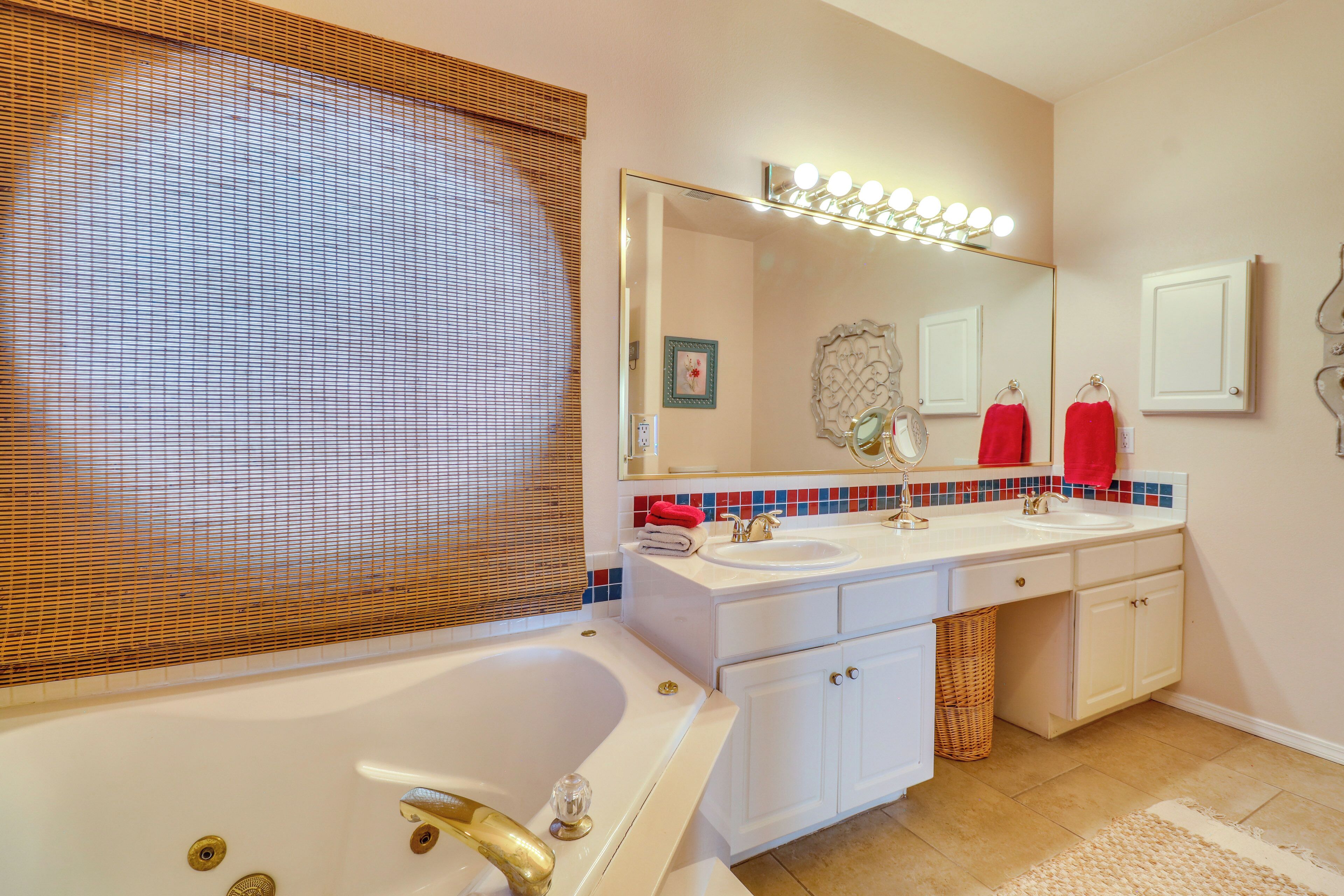 A bathroom with a bathtub, two sinks, a vanity with drawers, a large mirror with lights, and a decorative wall tile backsplash.