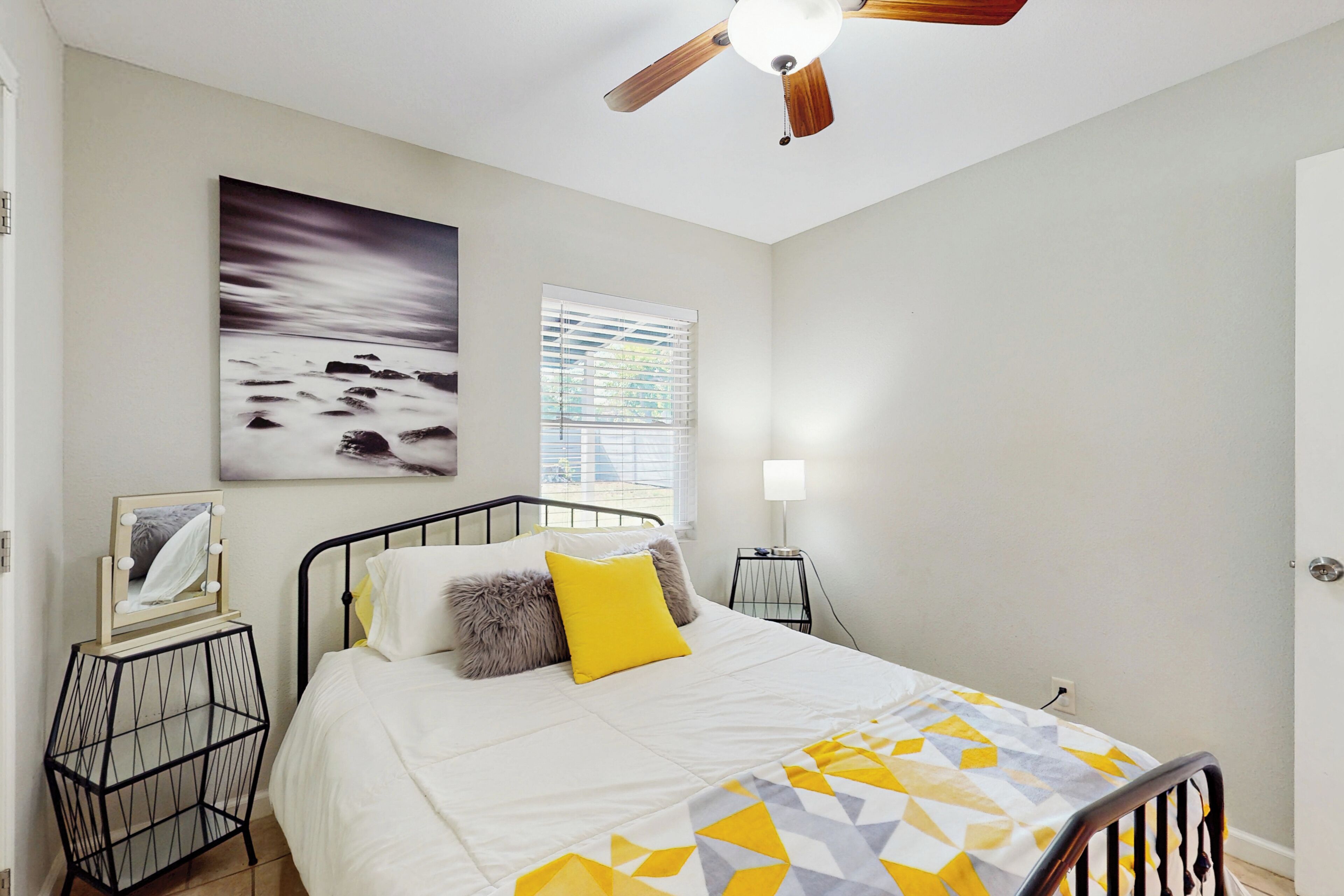 Ein Schlafzimmer mit Bett, Deckenventilator, Fenster mit Jalousien, Nachttisch mit Lampe und einem Wandbild.