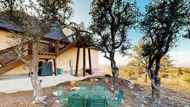 A rustic outdoor seating area with a metal table and chairs, surrounded by trees and a building with a sloped roof. - 'lone Star Leisure' Condo w/ Hill Country Views (Fredericksburg)