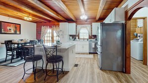 Una cocina moderna con vigas de madera, armarios blancos, un refrigerador de acero inoxidable y un comedor con sillas negras.