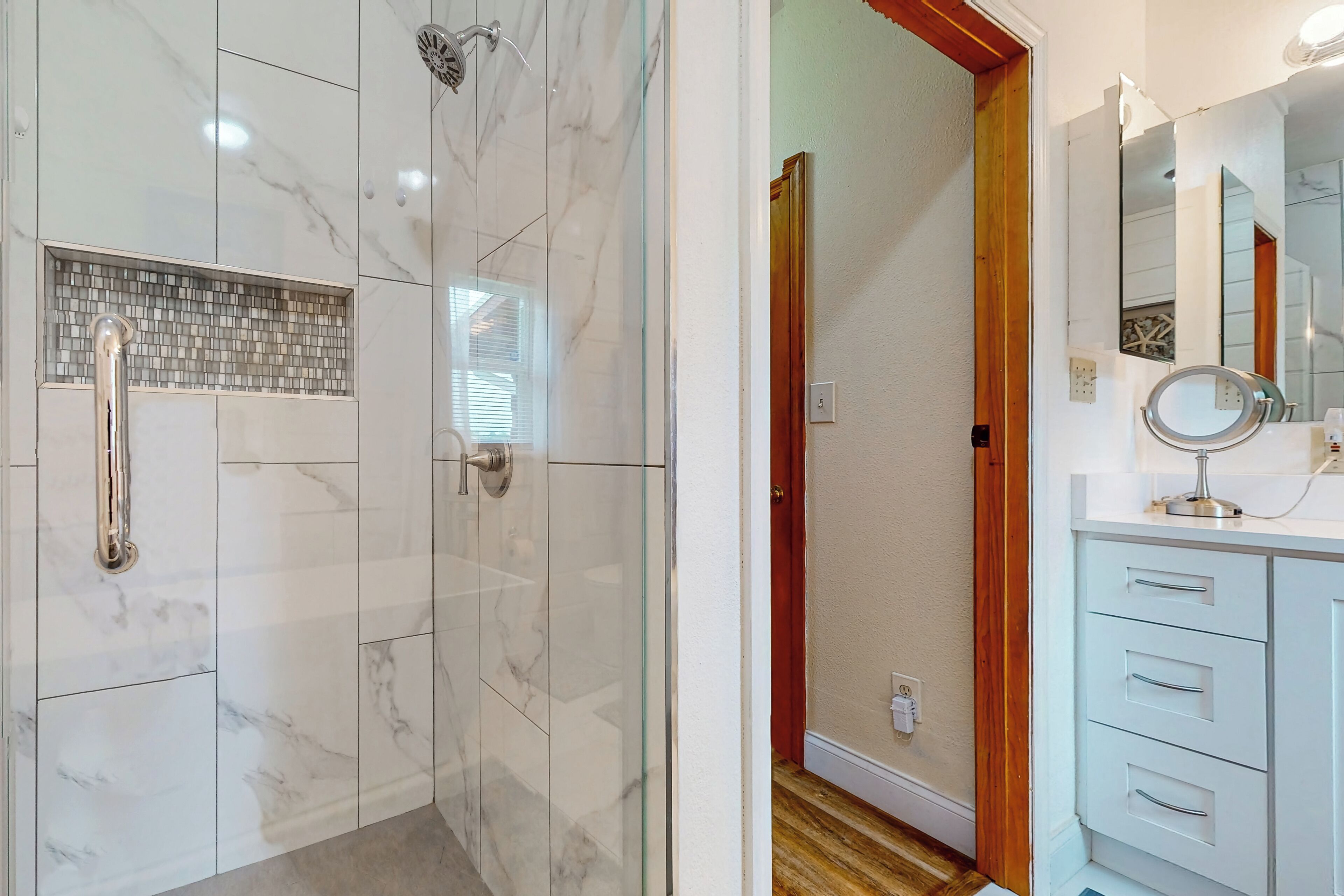 Une salle de bain dotée d’une douche à parois de verre, de carreaux de marbre blanc, d’une porte en bois et d’un meuble-lavabo blanc avec des tiroirs et un miroir.