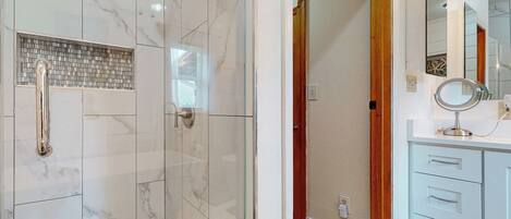 Une salle de bain dotée d’une douche à parois de verre, de carreaux de marbre blanc, d’une porte en bois et d’un meuble-lavabo blanc avec des tiroirs et un miroir.