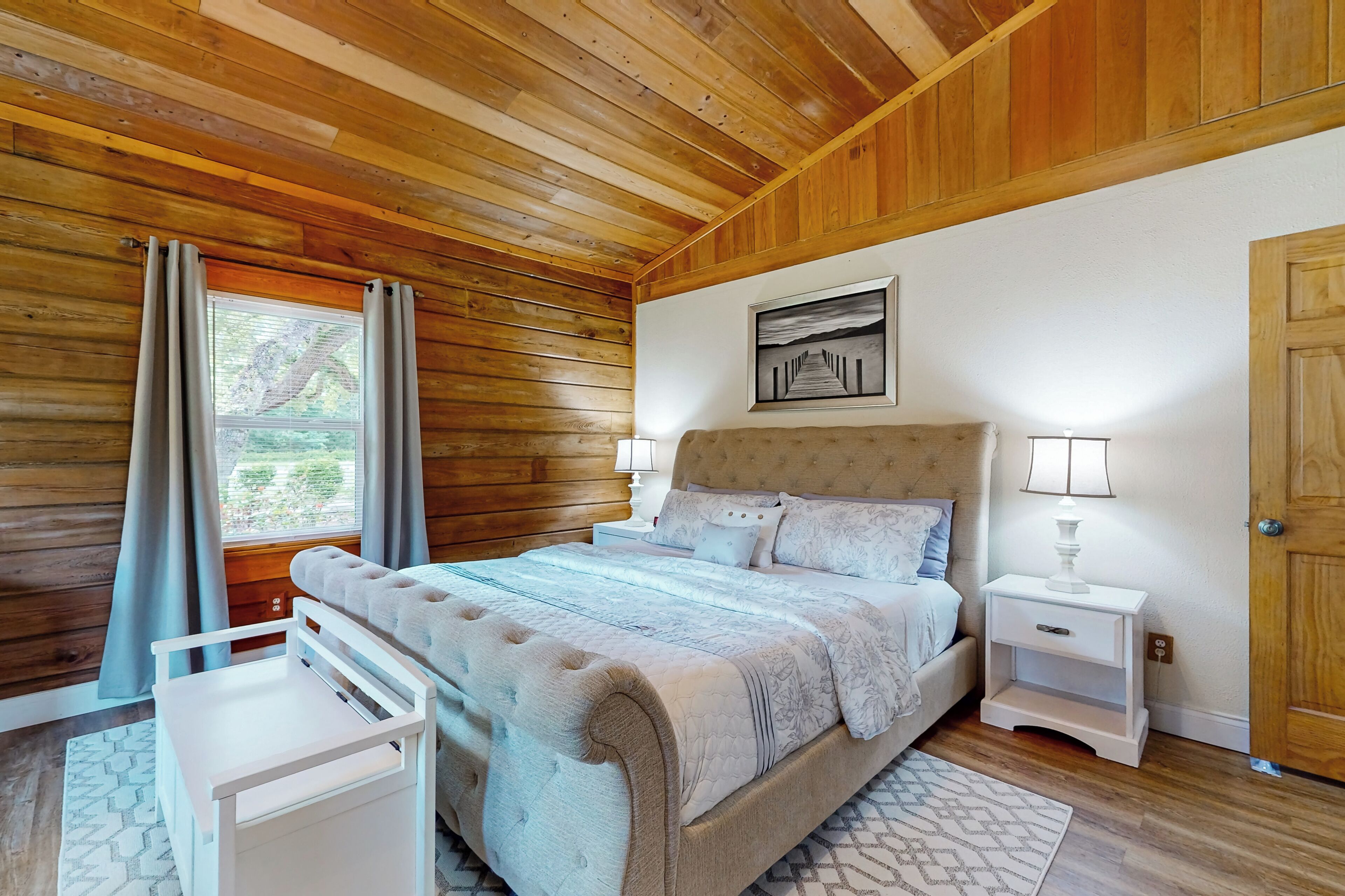 Une chambre avec un plafond en bois, un lit doté d’une tête de lit beige, deux tables de chevet avec des lampes et une fenêtre avec des rideaux.