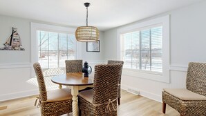 A dining area with wicker chairs, a round wooden table, and a hanging light fixture. - Walk to Downtown & Beach! Water-view Home (East Tawas)