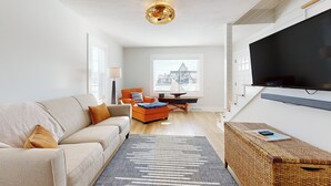A modern living room with a sofa, ottoman, and a television. - Walk to Downtown & Beach! Water-view Home (East Tawas)