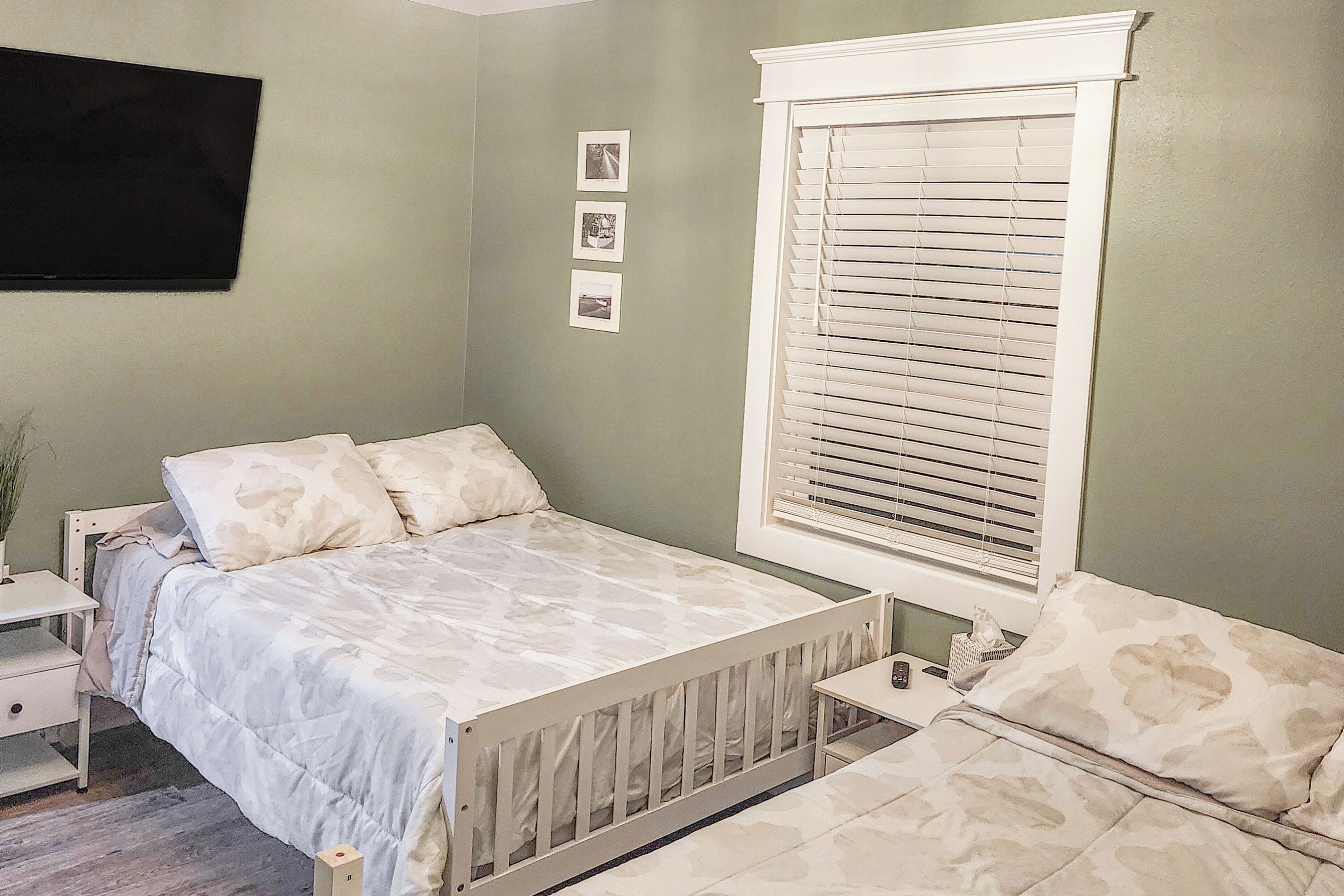 A bedroom with a bed, a nightstand, a bedside table, a window with blinds, and a wall-mounted TV.