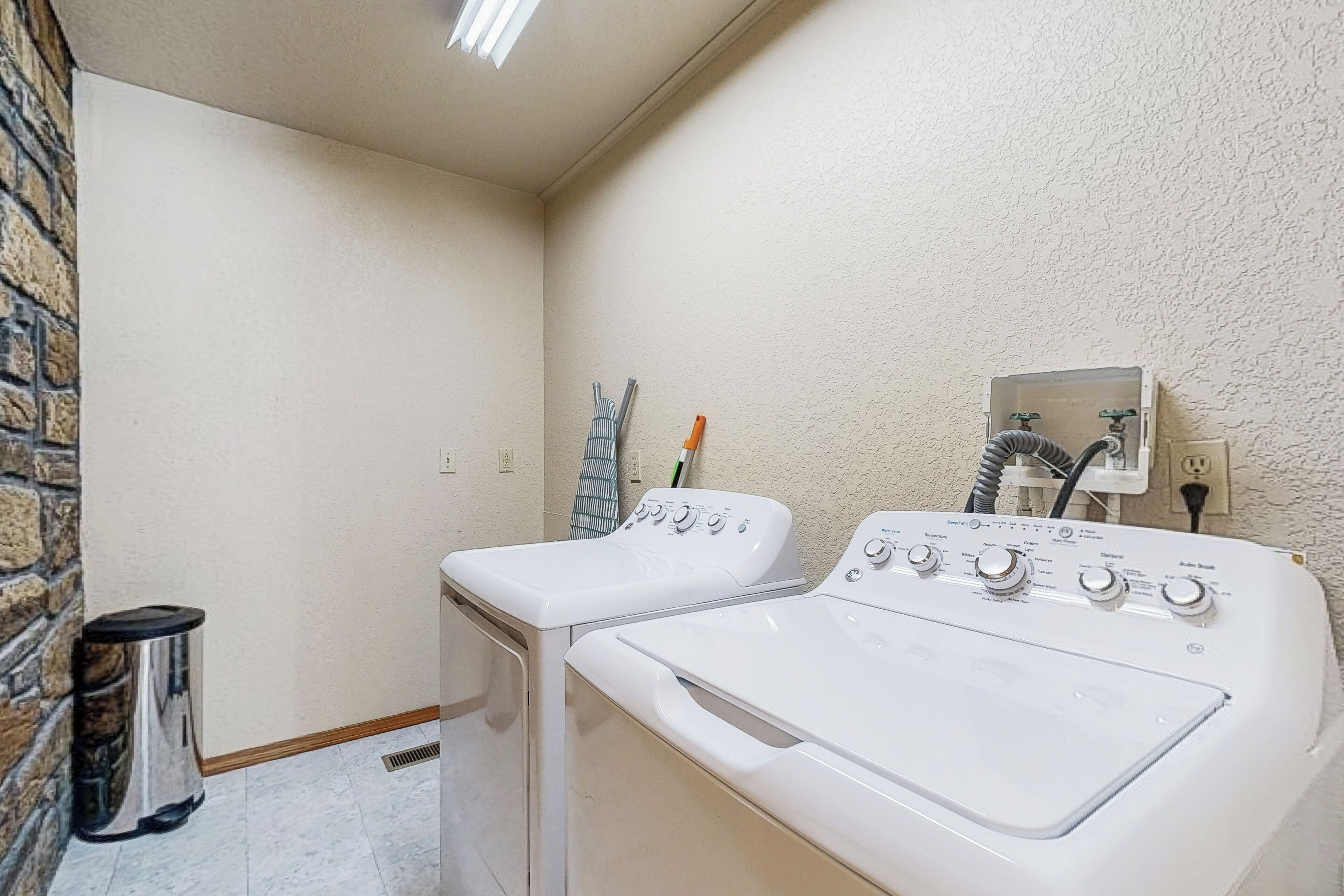 A laundry room with two white front-loading washing machines, a trash can, and a power outlet on the wall.
