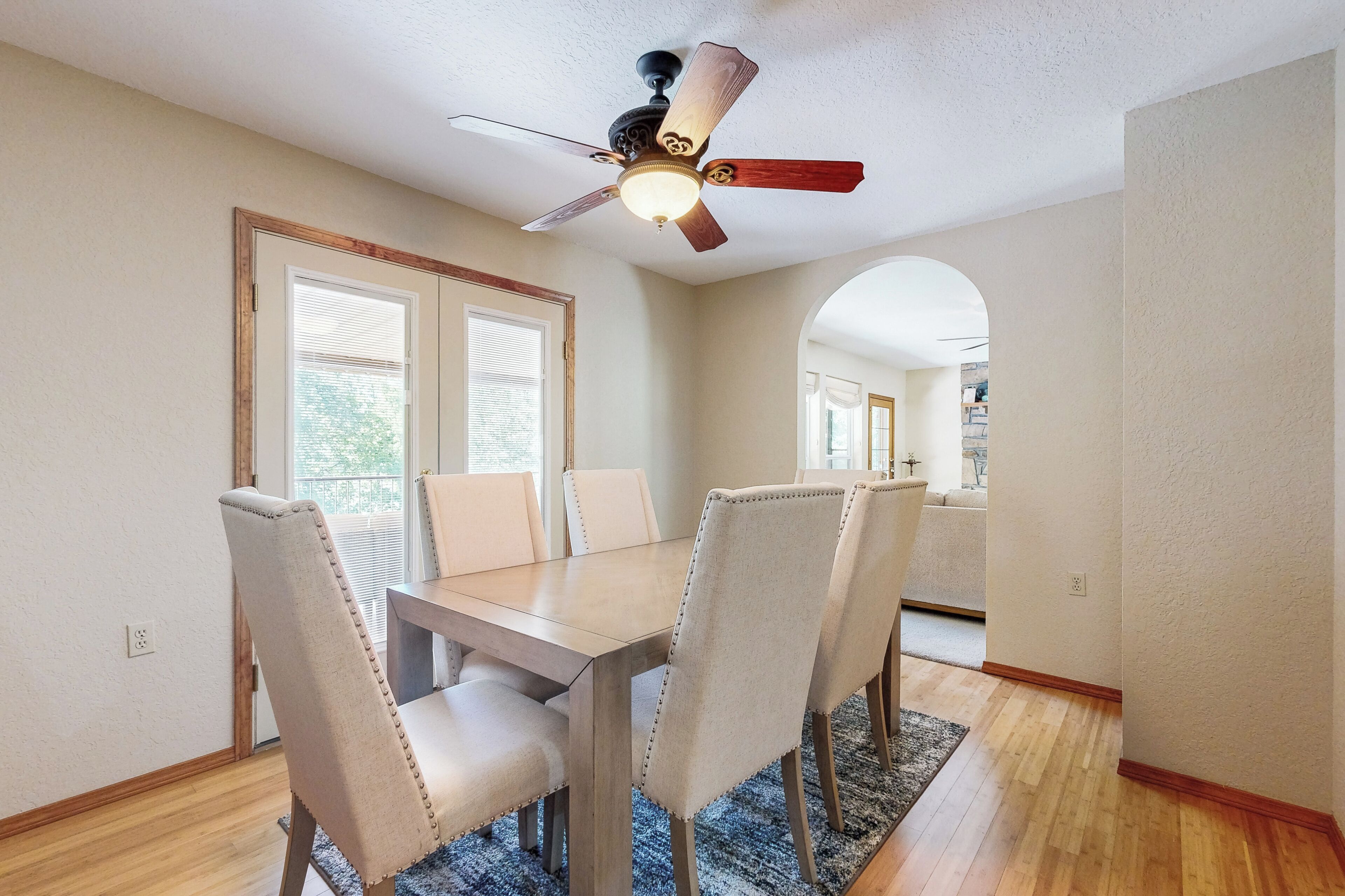 A dining area with a wooden table and beige upholstered chairs, a ceiling fan, and a view of a kitchen through an archway.
