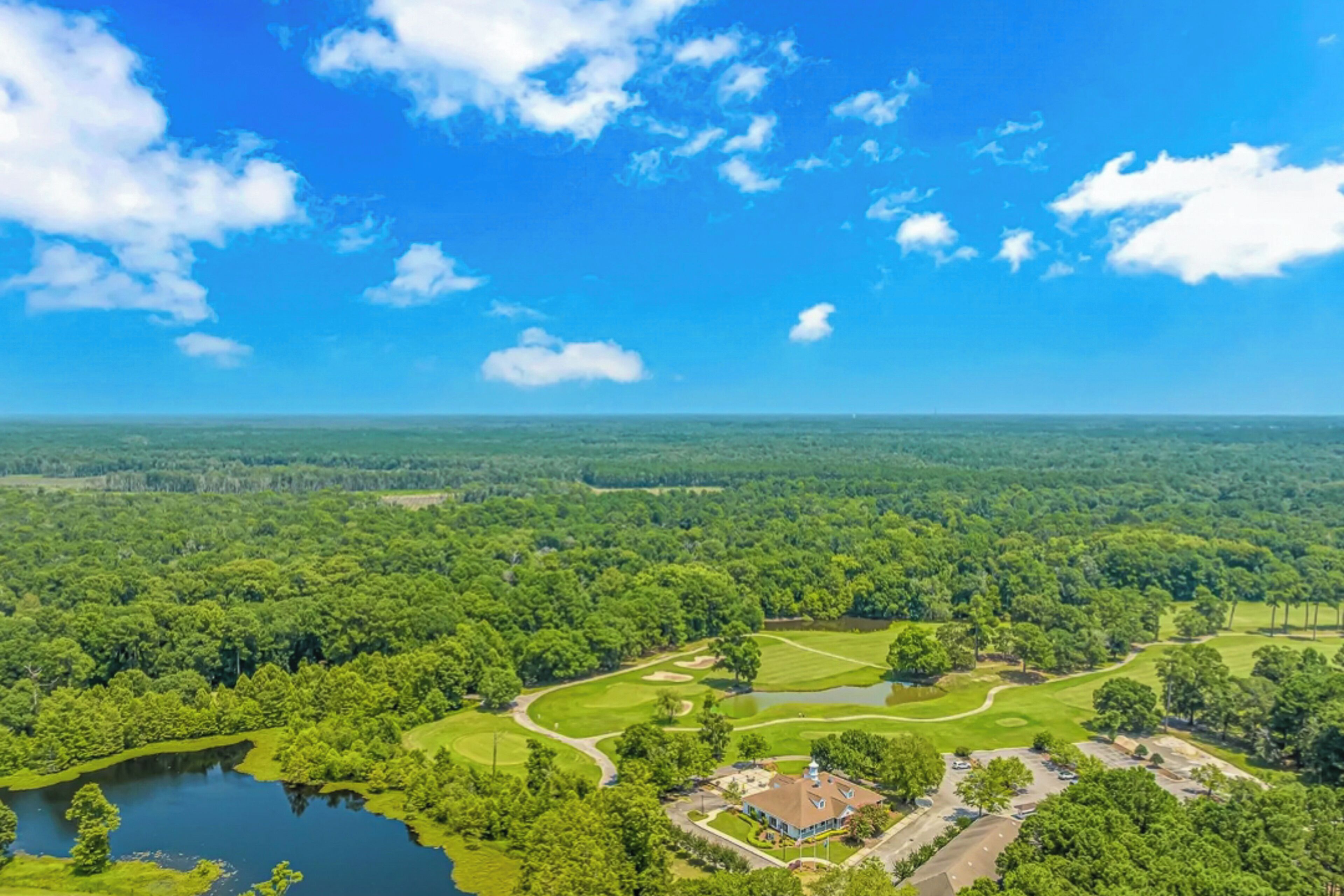 An aerial view of a golf course with a clubhouse, surrounded by dense forest and a clear blue sky.