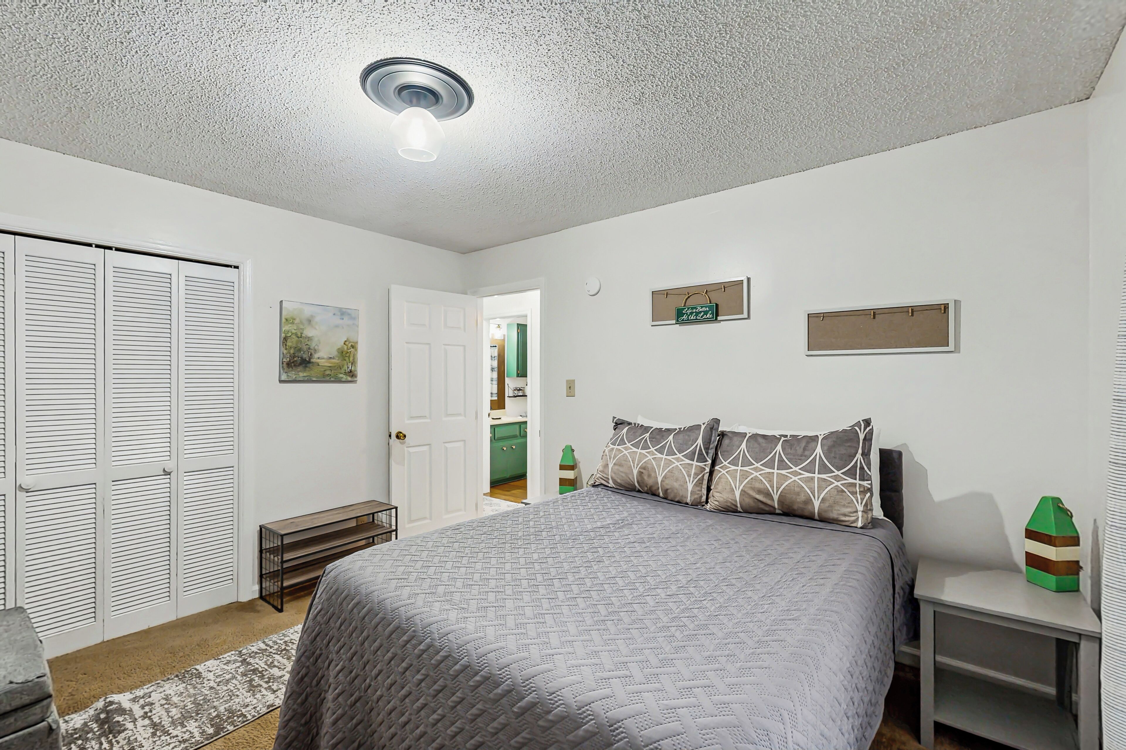 A bedroom with a bed, a bedside table, a bench, a closet, and a door leading to another room.