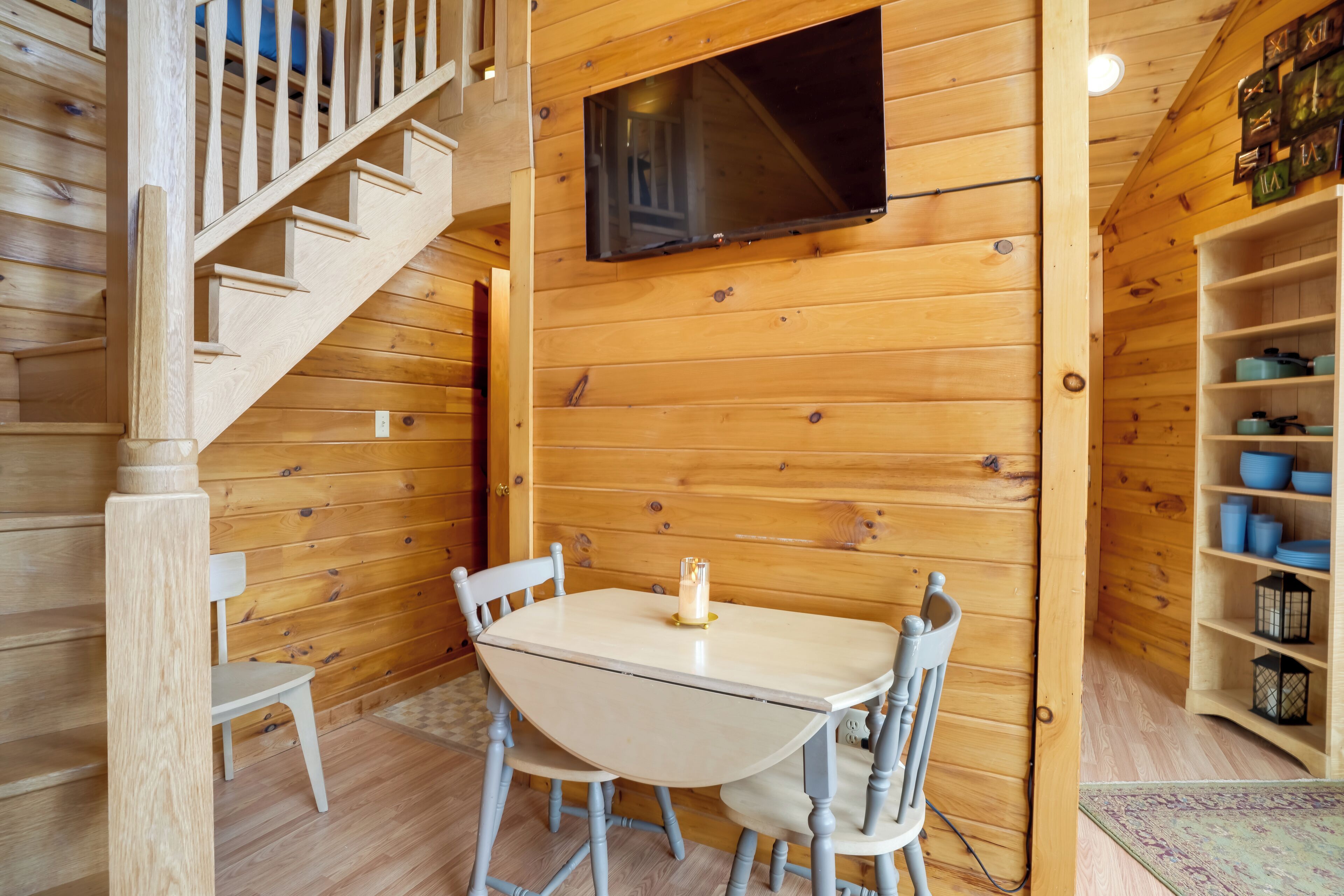 Un intérieur en bois avec un escalier, une table entourée de chaises, un téléviseur fixé au mur et une étagère avec des bols bleus.